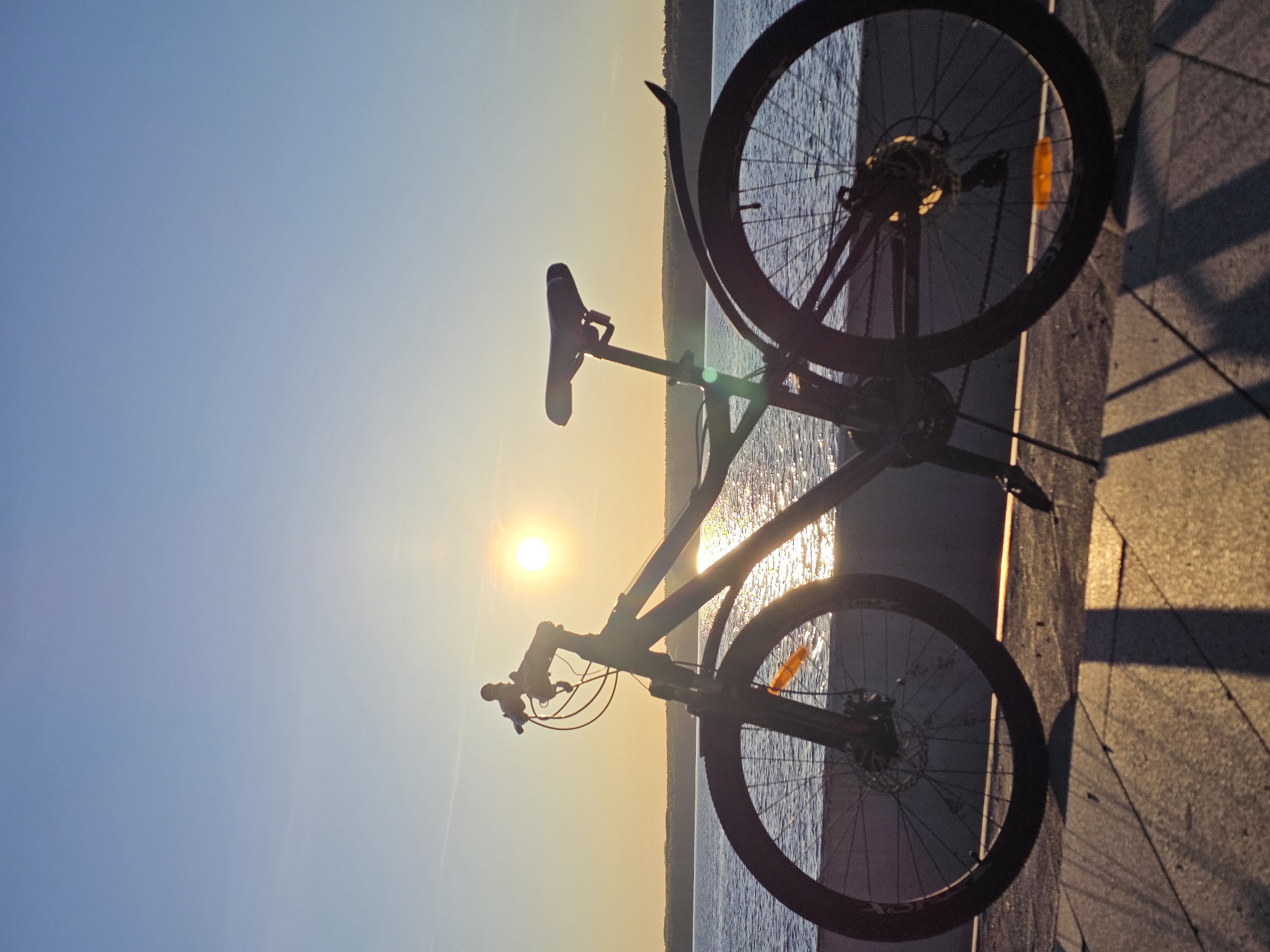 A bicycle rests against a concrete ledge with a body of water and a light-filled sky in the background. The bike is positioned vertically in the frame, casting a shadow on the concrete. The wheels are dark with orange reflectors. The seat and handlebars are silhouetted against the bright sky and water. The light appears to be from a sunset or sunrise. The location is Chaikovsky, Russia.