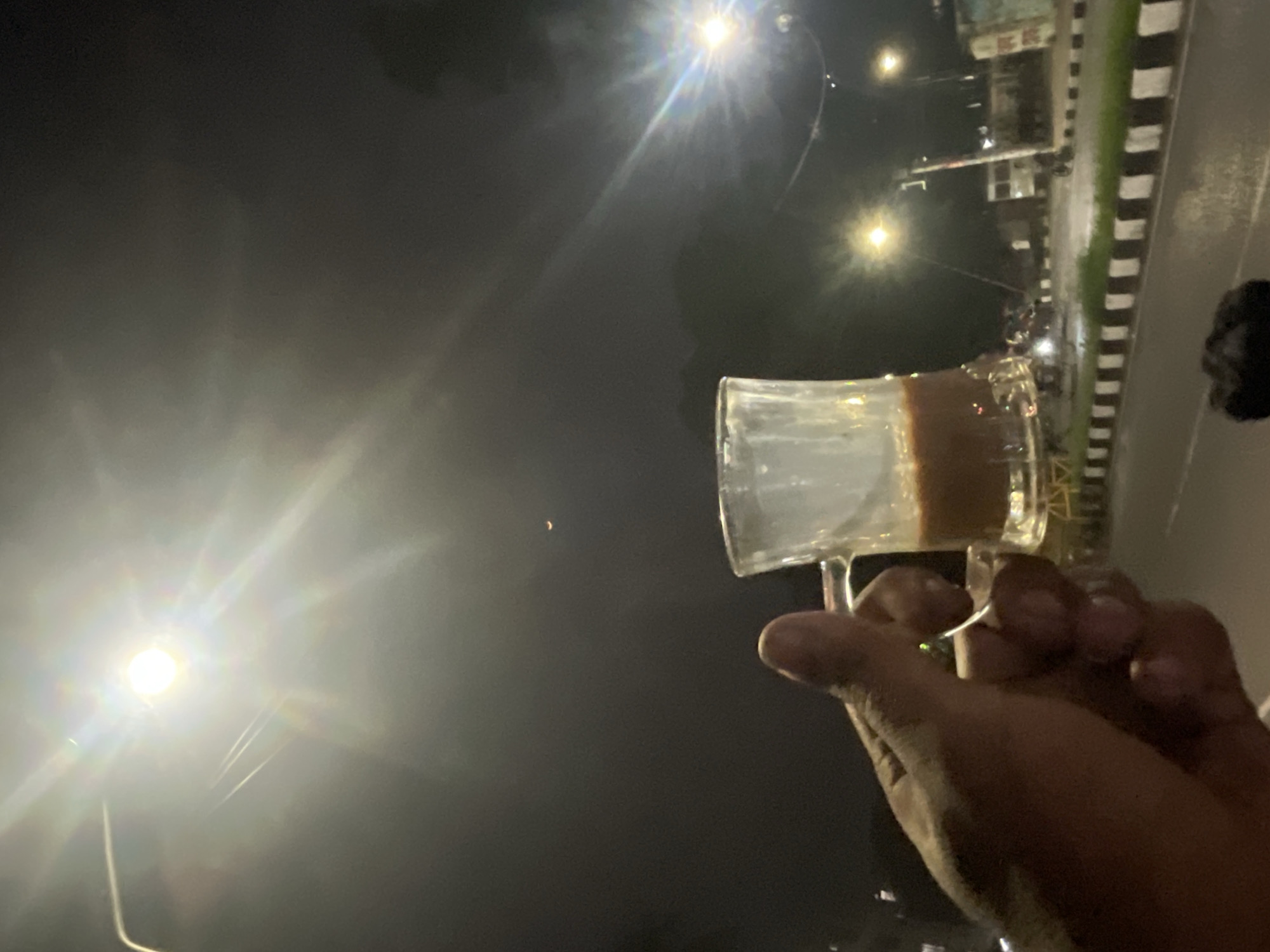 A person is holding a glass of tea in the dark, with streetlights illuminating the scene. A road with black and white striped edges is visible in the background. The image was taken in Dhaka, Bangladesh.