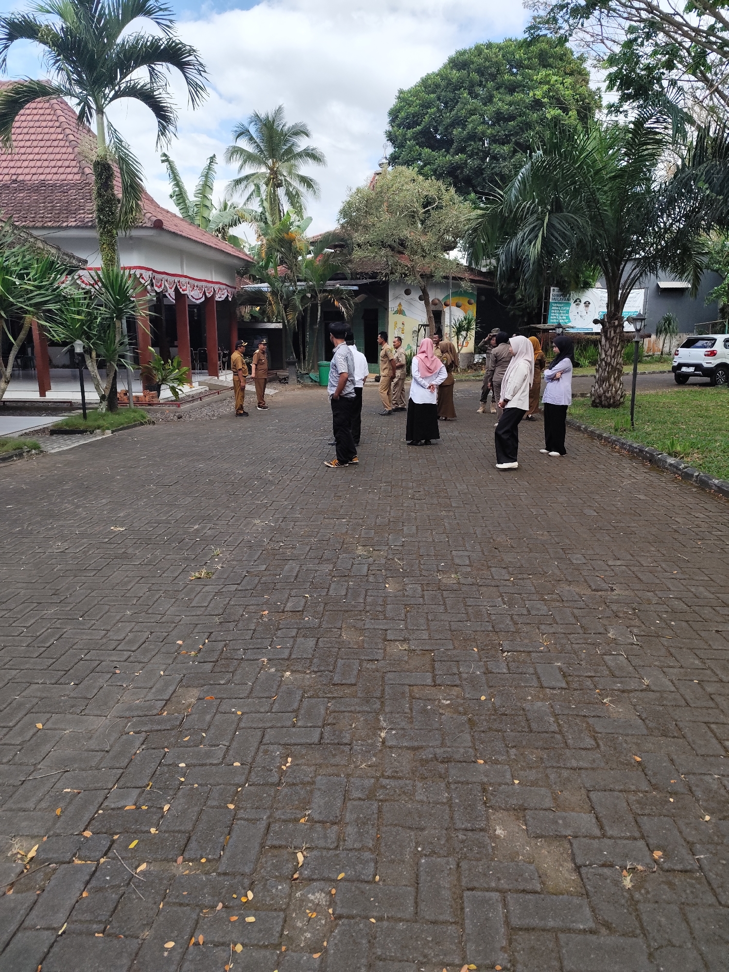 In Tegalarum, Indonesia, a group of people are gathered in an outdoor paved area in front of a building. The building has a distinctive red tiled roof and white walls decorated with a red and white banner. Several palm trees and other lush greenery surround the building and the open space. The paved area is made of dark interlocking bricks.
Among the group are several individuals dressed in light brown uniforms, suggesting they may be government officials or employees. Others are wearing casual attire, including women in headscarves. They appear to be engaged in a conversation or awaiting instructions, with some standing in small groups and others observing.
In the background, a white car is parked on the side of the road near a grassy area. The sky is partly cloudy, indicating an overcast day. The overall scene suggests a formal or semi-formal gathering outdoors in a tropical or sub-tropical environment.