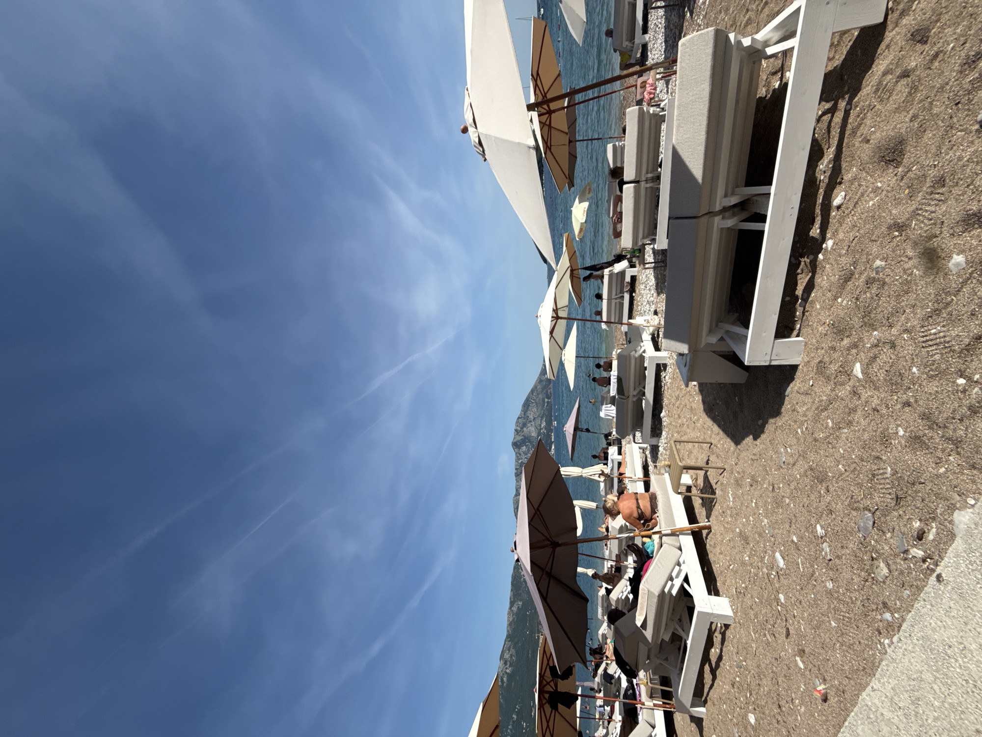 This image depicts a vibrant beach scene in Budva, Montenegro, under a clear blue sky crisscrossed by multiple faint contrails. In the distant background, calm blue sea waters meet a rugged coastline dominated by mountains. Several small boats are visible on the water.
The midground and foreground are occupied by a resort-style beach setup. Numerous sun loungers with white wooden frames and light grey cushions are arranged in rows across the sand and pebble beach. Many of these loungers are occupied by individuals relaxing. Large beach umbrellas, predominantly light cream or beige in color, are strategically placed to provide shade over the loungers.
Specific observable subjects include:
- A woman in a leopard-print bikini top and dark bottoms sits on a sun lounger in the lower center, facing away from the viewer towards the sea.
- To her left, partially obscured by an umbrella, a person in dark attire reclines on a lounger.
- In the far right foreground, two unoccupied sun loungers with light grey cushions and white frames are prominently visible.
- Small, dark-colored side tables are situated between some loungers, one holding a drink cup.
- The beach surface consists of light brown sand mixed with numerous small, light-colored pebbles and a few larger stones.
- A concrete ledge or seawall is visible along the bottom left edge of the frame.
- In the distance, closer to the water, more people are visible, some standing in the shallow water.
The scene conveys a sunny, leisure-oriented atmosphere. Shadows cast by the umbrellas and loungers indicate a clear day with direct sunlight.