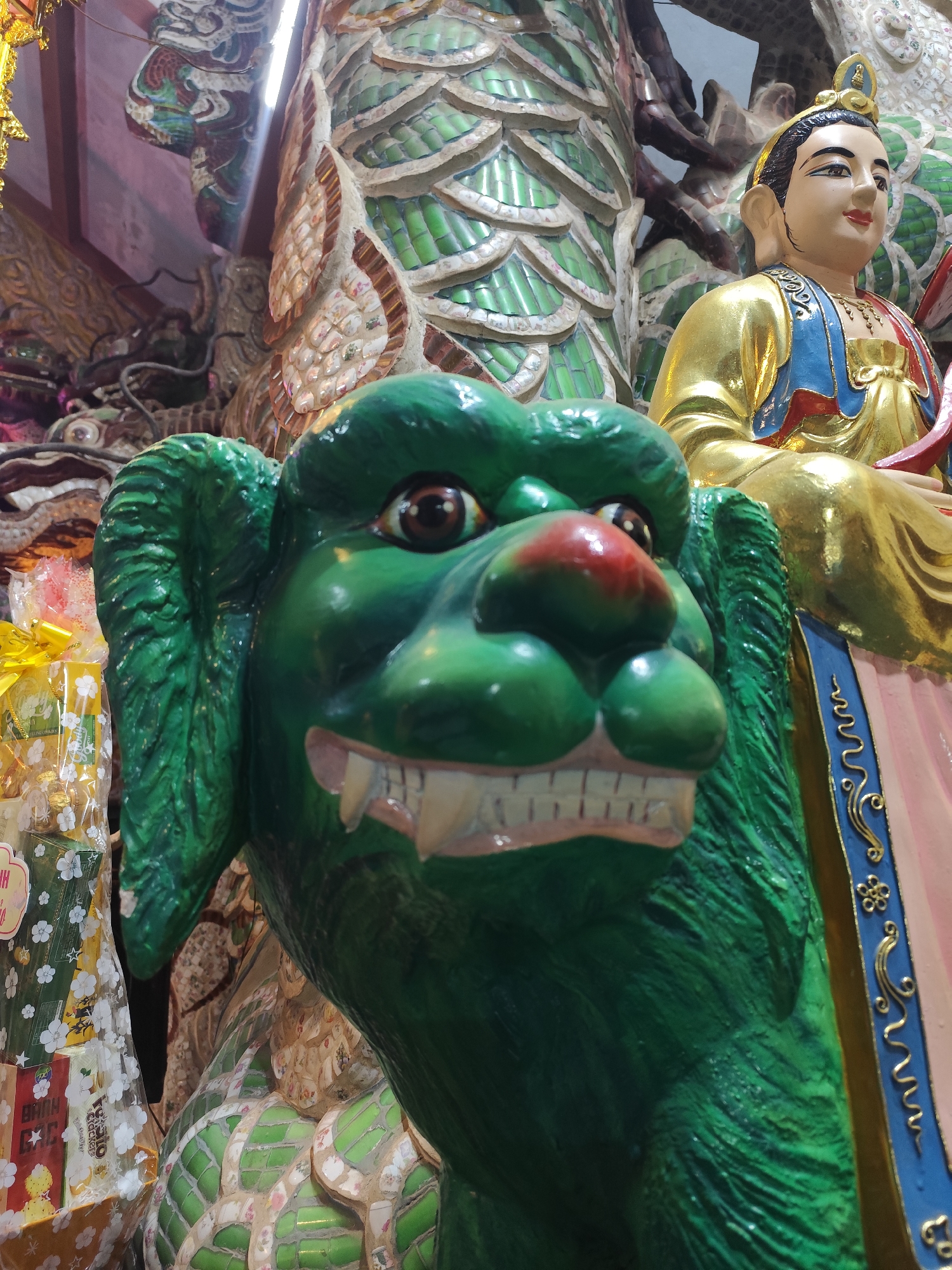 The image shows a close-up of a statue of a green dragon-like creature in front of a statue of a person wearing a gold and blue robe. The dragon has a red nose, large brown eyes, and sharp white teeth. The person is seated and appears to be holding something in their lap. The background is blurred, but it appears to be an indoor setting with decorative elements. The statue is likely located in a temple or shrine in Lâm Viên - Đà Lạt, Vietnam, given the style and religious iconography. There are also some gift baskets wrapped in clear plastic with yellow bows visible on the left side of the image. The overall impression is one of vibrant religious or cultural art.