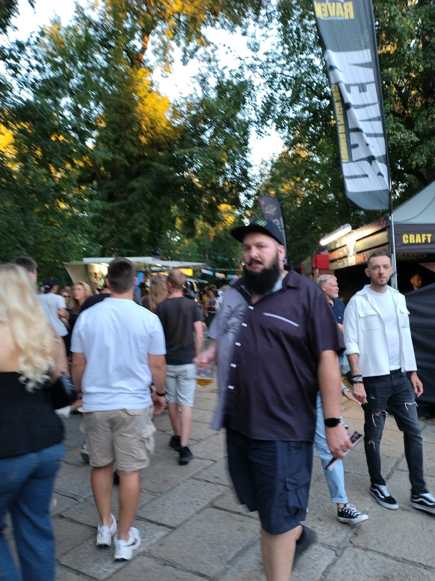 The image depicts a bustling outdoor scene at an event, likely a festival or market, with numerous people visible. In the foreground, a man with a beard and wearing a dark, patterned shirt and dark shorts is the most prominent figure. To his right, another man in a white shirt and distressed black jeans is also clearly visible. Behind them, other individuals are walking and interacting. The environment includes trees, tents, and what appear to be food or vendor stalls in the background. The ground is paved with flagstones. A tall, vertical banner with the word "RAVEN" is visible on the right side of the image, suggesting a branding or sponsorship presence. The overall atmosphere suggests a daytime event with moderate activity. The image was taken in Písek, Czechia.