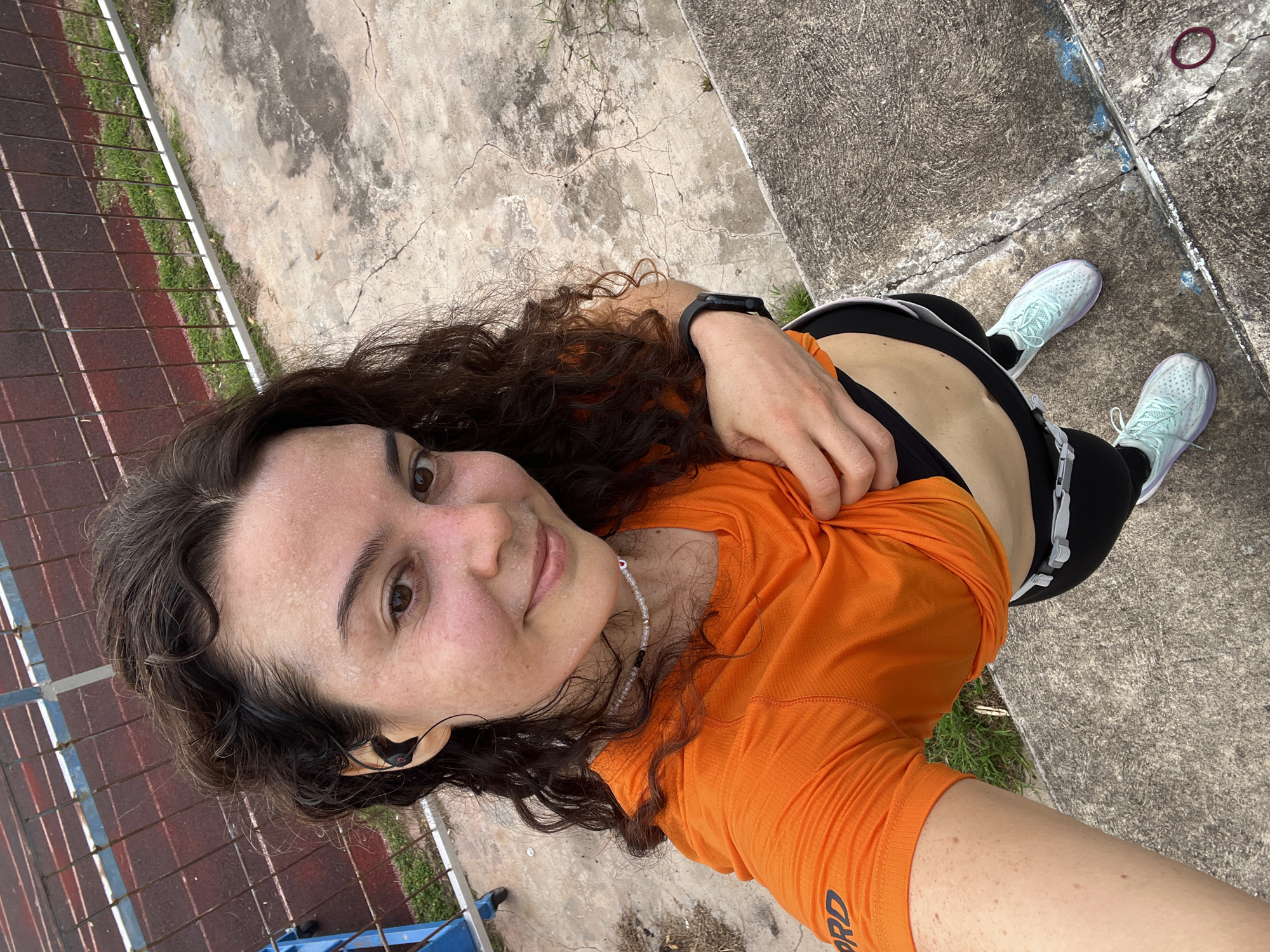 A young woman with curly brown hair is taking a selfie outdoors. She is wearing an orange t-shirt, black athletic shorts, and light blue sneakers. Her face is flushed and appears sweaty, suggesting she has been physically active. She is wearing black earbuds and a thin necklace. The background shows a running track with a metal fence and concrete steps. The location is identified as Phan Thiết, Vietnam.