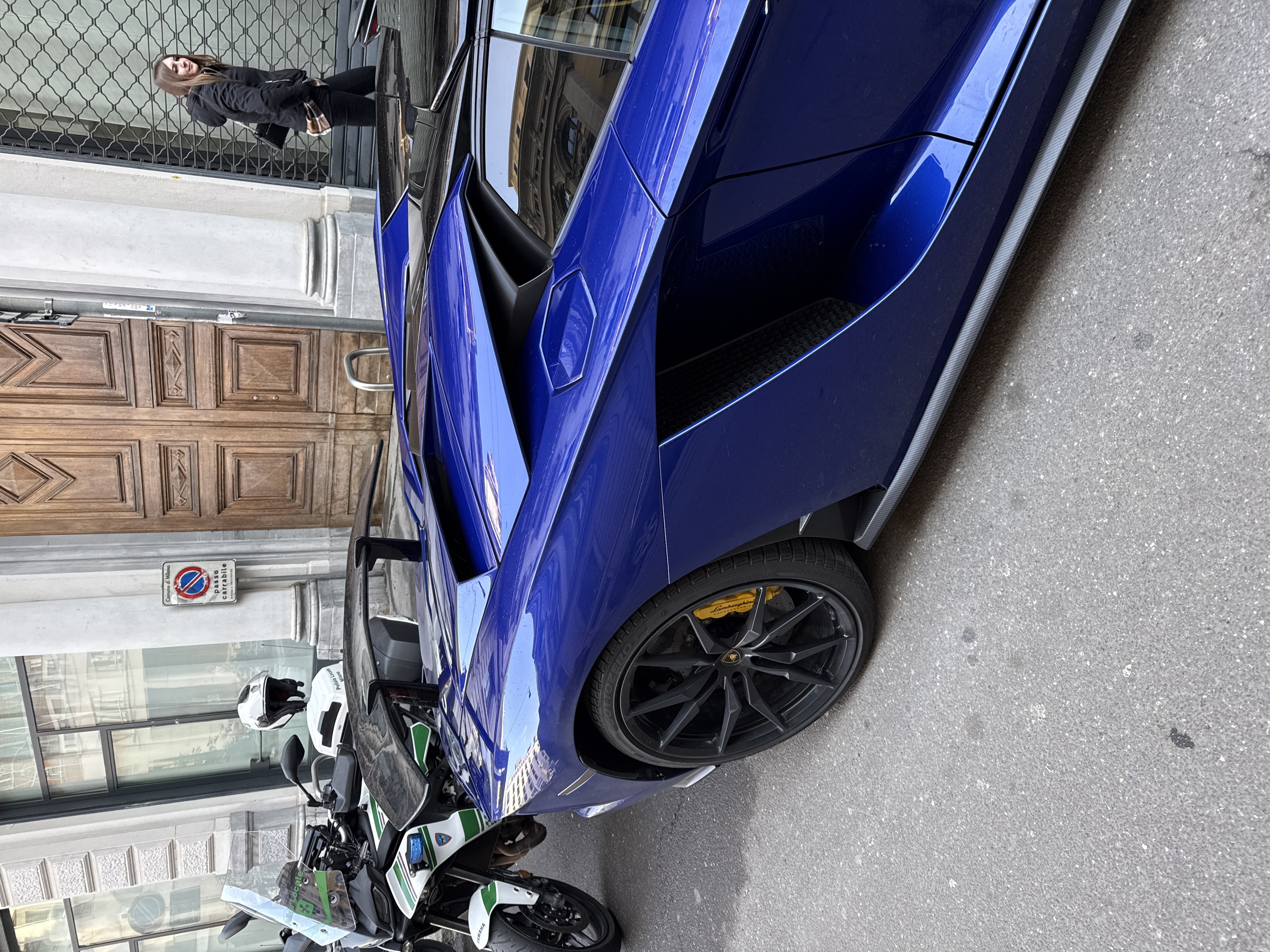 The image depicts an urban street scene in Milan, Italy, confirmed by "Comune di Milano" and "Polizia Locale Milano" signage. A dark blue Lamborghini supercar is prominently featured on the right, parked parallel to the curb, displaying a black multi-spoke wheel with a yellow Lamborghini-branded brake caliper and a rear spoiler. To the left of the supercar, a white and green Yamaha motorcycle, identified as a "Polizia Locale Milano" vehicle, is parked. A white police helmet rests on the motorcycle's rear. In the background, a light-colored building with large windows reflects its surroundings. Adjacent to the building, a "Comune di Milano passo carrabile" (no parking) sign is visible near a large, ornate wooden double door. On the far right, a female pedestrian with brown hair, wearing a black hooded jacket over a plaid shirt and carrying a black shoulder bag, walks away from the camera past a building section with a metal security grate. The street surface is asphalt.