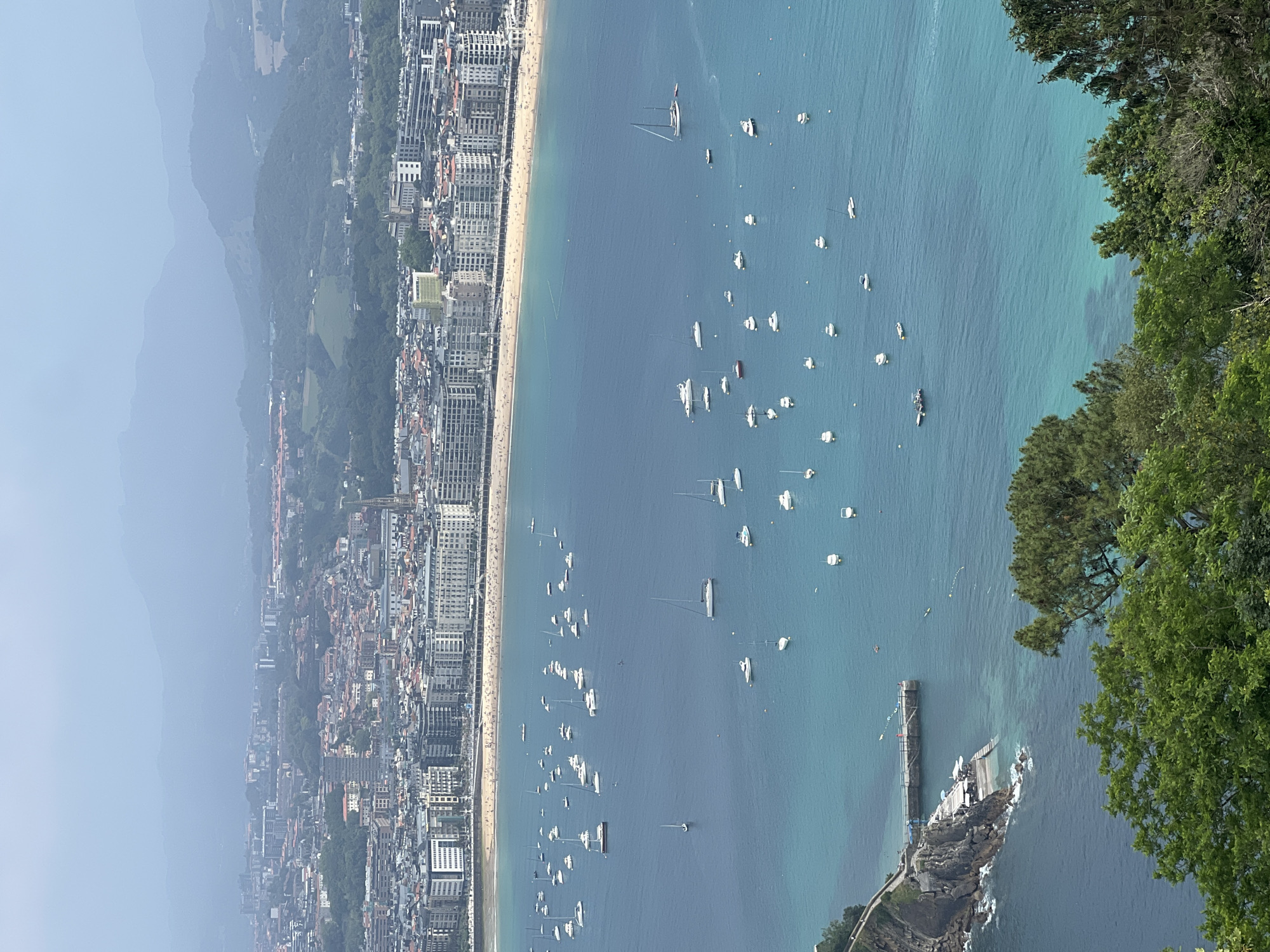The image is an elevated, wide-angle view of La Concha Bay in San Sebastián, Spain, on a hazy day. The bay is filled with numerous boats of varying sizes, mostly sailboats and smaller motorboats, scattered across the turquoise and deep blue water. In the background, a densely populated city lines the curved coastline, with buildings of varying heights and architectural styles. Beyond the city, rolling green hills and mountains are visible, partially obscured by atmospheric haze.
In the foreground, lush green trees frame the bottom of the image. To the left, a rocky outcropping juts into the water, with a small pier or walkway extending from it, featuring umbrellas and seating areas on a terrace. A few small boats are moored near the rocky area. The beach, populated by many sunbathers, is clearly visible along the city's waterfront. The water exhibits a gradient of color, from a bright, clear turquoise near the shore to a deeper blue further out in the bay. The sky is overcast and pale blue, contributing to the hazy effect.