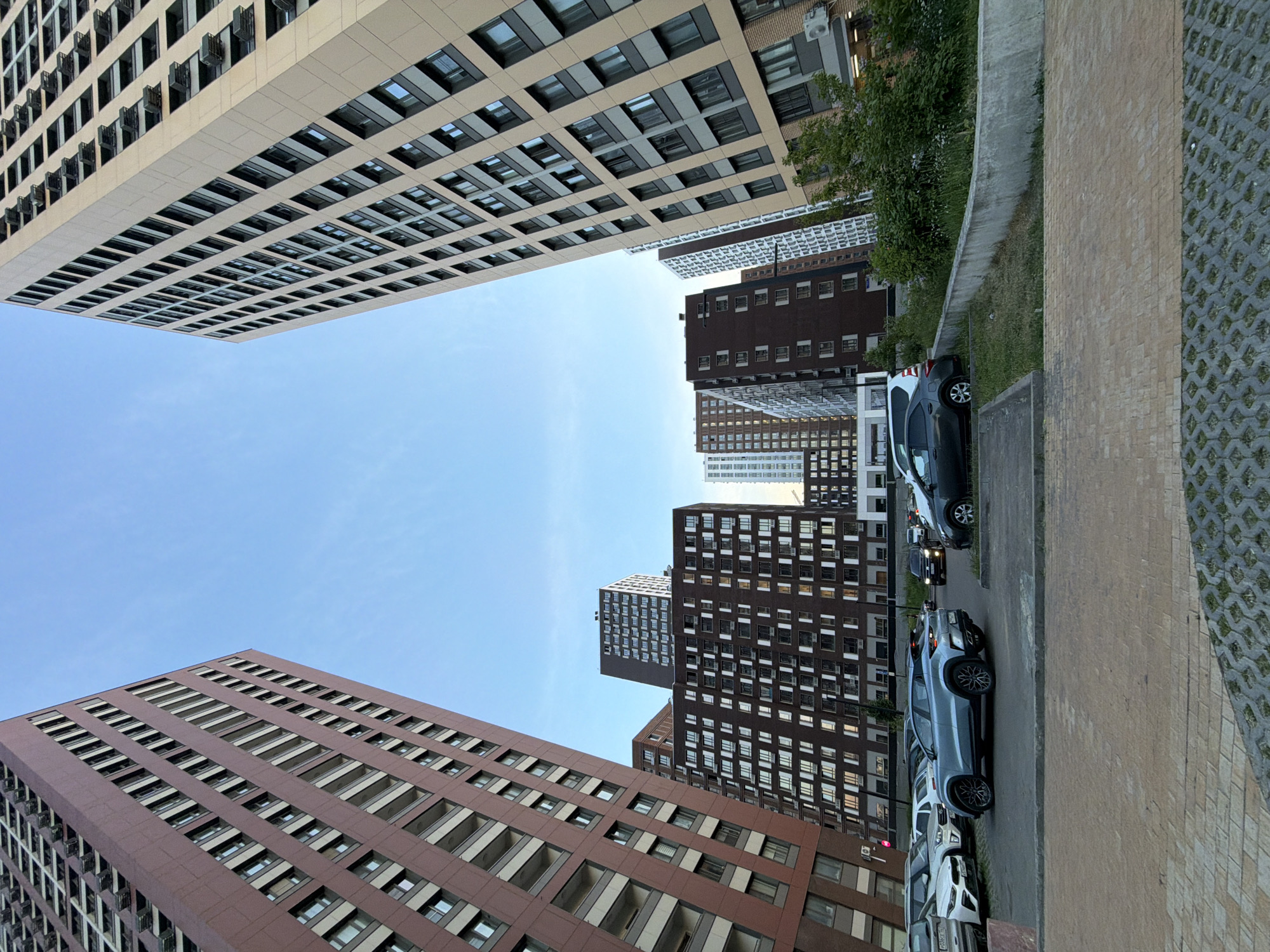 In the image, a modern residential complex is depicted in Moscow, Russia. Two tall, multi-story buildings dominate the frame, one on the left with a reddish-brown facade and balconies, and another on the right with a light beige facade and a repeating pattern of windows and dark accents. Several other buildings of varying heights and architectural styles are visible in the background, creating a dense urban landscape.
In the foreground, a parking lot is situated in front of the buildings. Several cars are parked, including a light blue SUV in the center, a white sedan and another white vehicle to its left, and a dark gray sedan to the right. The ground in the parking area is paved with gray concrete blocks, with some green vegetation and a curved concrete planter box visible on the right side. The sky is clear and blue. The scene appears to be a residential area within a city.