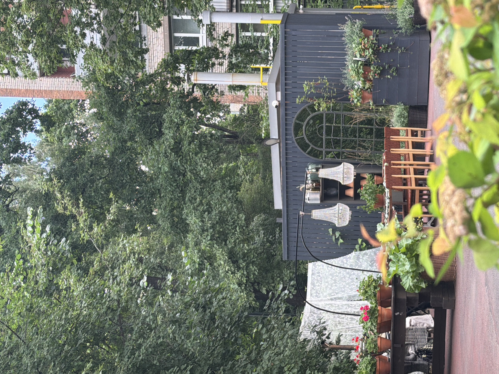 This image captures an outdoor seating area with a dark wooden fence and a large arched window. Several decorative chandeliers hang from the fence. Wooden chairs are neatly stacked to the right of the fence. Potted plants are arranged on shelves and in planters along the fence line, with red flowers and lush green foliage visible. A white translucent canopy is set up in front of the fence. In the background, a brick building with windows can be seen through dense green trees. The scene appears to be set during the daytime, with soft lighting suggesting it might be overcast or late afternoon. The overall impression is one of a well-maintained, charming outdoor space, possibly a cafe or restaurant terrace. There is no visible text in the image.
