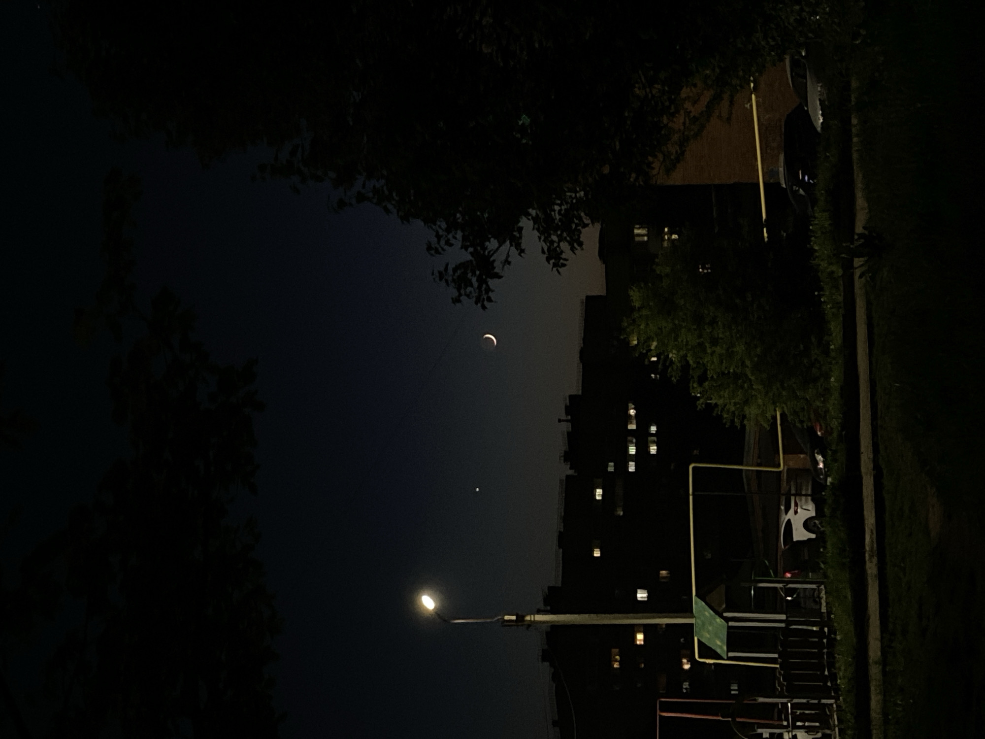 The image depicts a nighttime scene in Votkinsk, Russia, framed by trees at the top and left, and a building on the right. A crescent moon is visible in the dark sky, along with a dim light source below and to the left of it, possibly a star or planet. Below the moon is a tall apartment building with lit windows. A street light illuminates a playground in the foreground. A car is parked near the building.