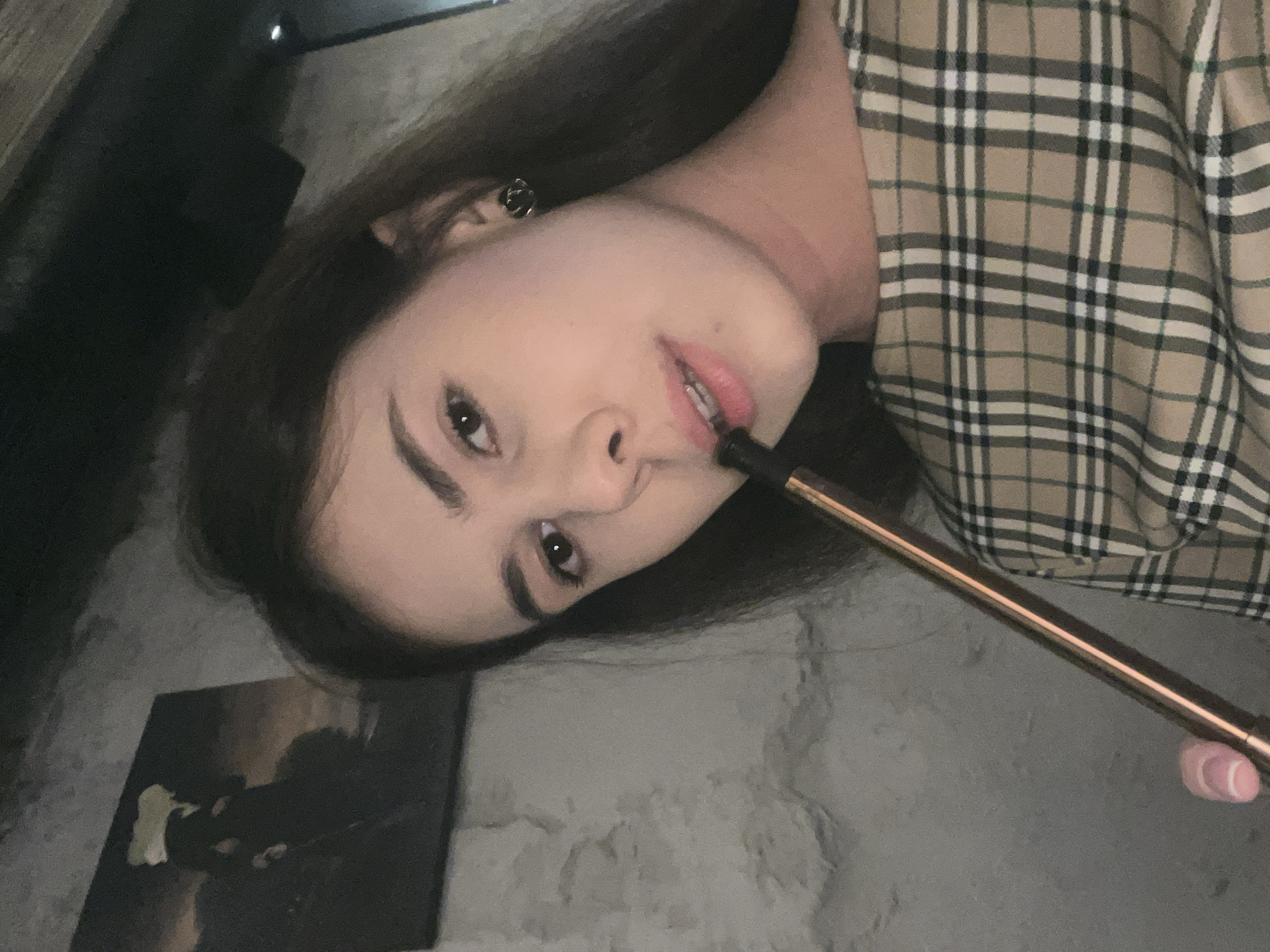 A young woman is depicted in a close-up, chest-up shot in an indoor setting in Unknown, Unknown. She has long, dark brown hair, dark eyes, and light skin, and is looking directly at the camera with a neutral expression. She wears a small, dark, possibly floral or knot-shaped stud earring in her visible right ear. Her attire consists of a beige/tan plaid top featuring a grid pattern of black, white, and thin reddish-brown lines. She holds a slender, metallic (rose gold/copper-toned) hookah hose mouthpiece to her lips, with a black tip inserted into her mouth. Her left thumb, displaying a French-tipped nail, is visible holding the hose. The background features a textured, light gray wall. A dark-toned framed picture, depicting silhouetted figures in a landscape with a lighter element above them, hangs on the wall behind her left shoulder. The upper portion of the image shows a dark, indistinct ceiling or upper wall. The scene is dimly lit.