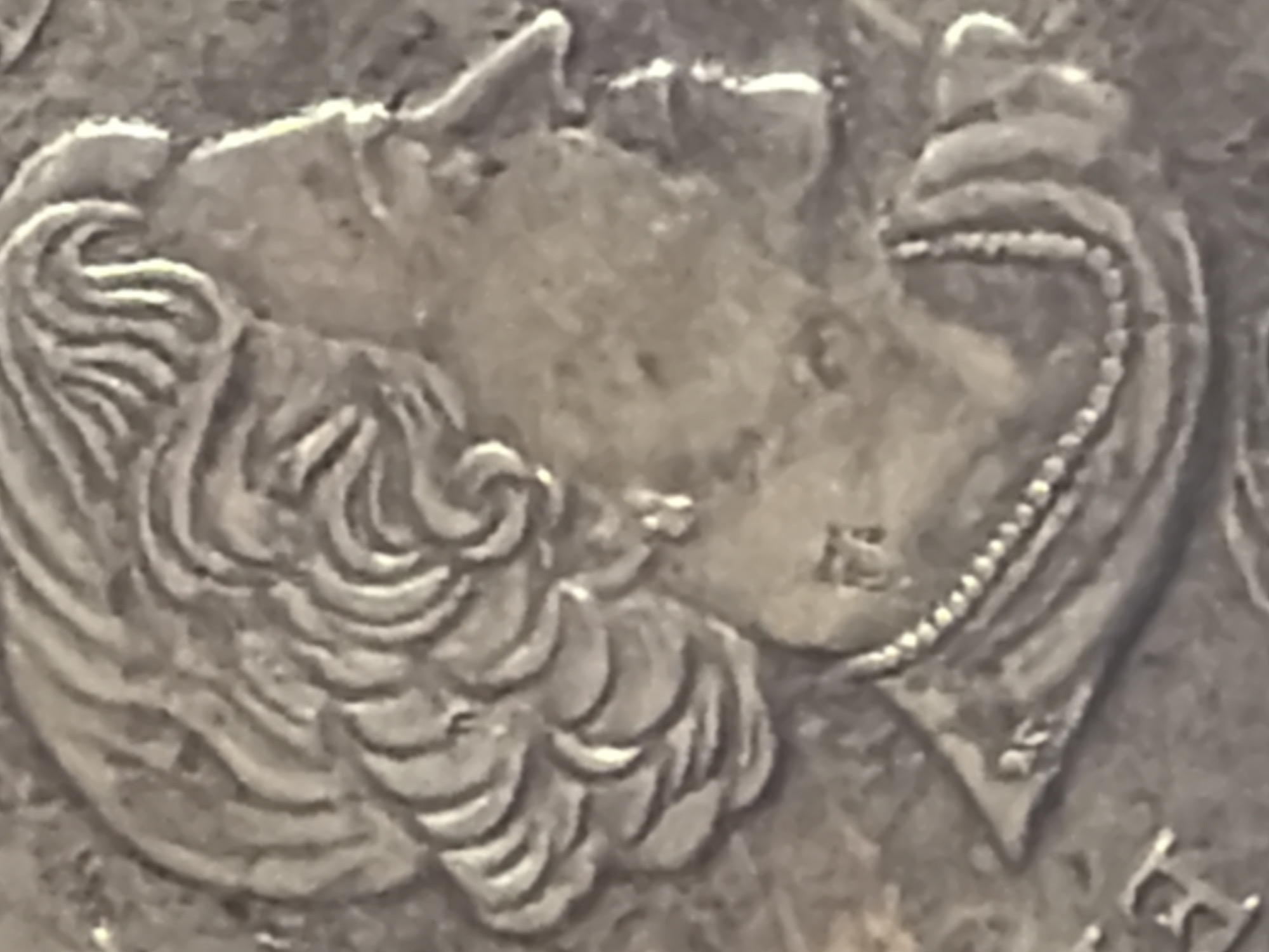 The image is a close-up, grayscale photograph of a coin, likely a commemorative one, depicting a profile of a person, possibly a monarch, facing right. The individual has curly hair, a prominent nose, and is wearing an earring and a beaded necklace. The relief is detailed, with clear lines defining the facial features, hair texture, and jewelry. There appear to be some small markings or inscriptions on the coin near the subject's neck and shoulder area, but they are not clearly legible. The coin itself has a metallic sheen, indicating it's made of a silver-toned metal. The overall impression is of a finely crafted piece of currency or collectible. The image does not contain any information about the city of Arcahueja, Spain.