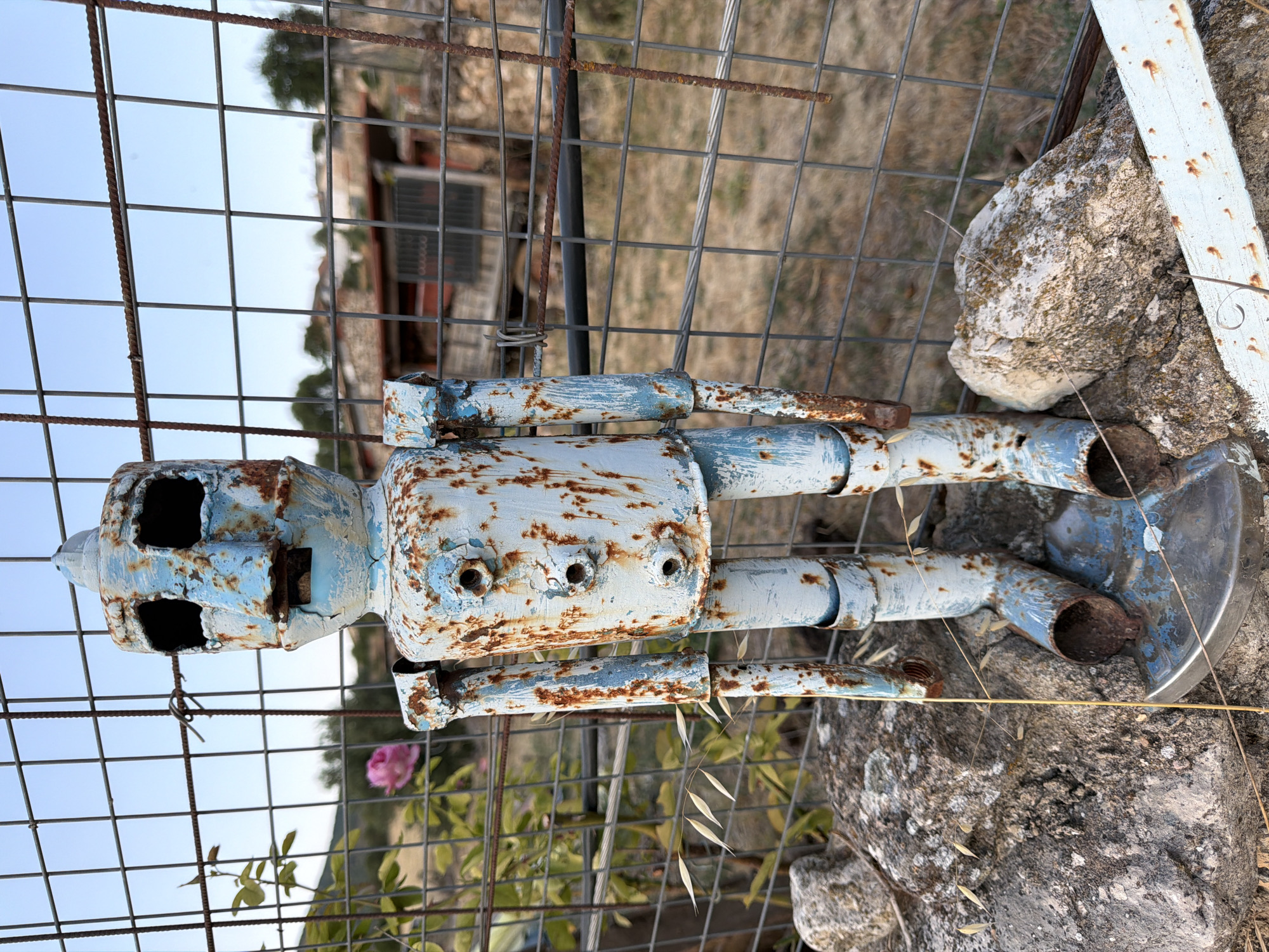 A weathered, pale blue and rusty metal robot sculpture stands in front of a wire fence in Salmerón, Spain. The robot has a cylindrical body with three visible bolt-like circles on its chest and articulated arms and legs made from pipes. Its head is blocky with large, dark rectangular eye sockets and a small, rectangular mouth. The robot is positioned on a metal circular base that rests on two large rocks. Behind the robot, the wire fence is visible, with vegetation and buildings in the background under a clear sky. A pink rose is blooming on a vine to the left of the robot, partially obscured by the fence. The environment appears to be outdoors, possibly in a rural or semi-rural setting within the town.