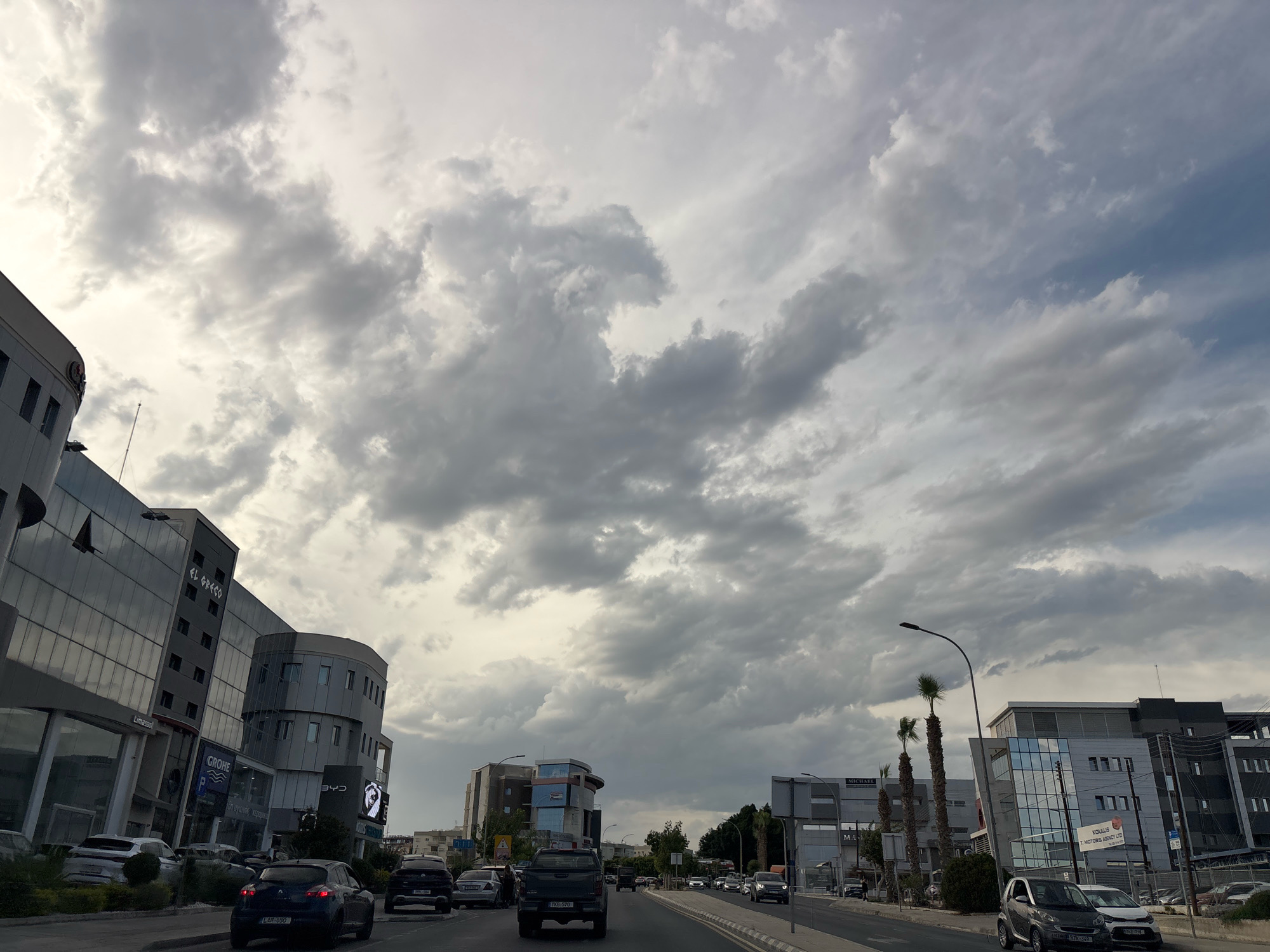 The image depicts a cloudy sky over a city street in Agios Athanasios Municipality, Cyprus. Modern buildings line the right side of the street, featuring reflective glass facades and architectural elements like curved sections and balconies. Palm trees are visible along the street, interspersed with other foliage. Several vehicles are driving on the road, including cars and a pickup truck. The sky is filled with dramatic, grey clouds, suggesting overcast or potentially rainy weather. The overall impression is a view from a vehicle traveling down a city road on a cloudy day.