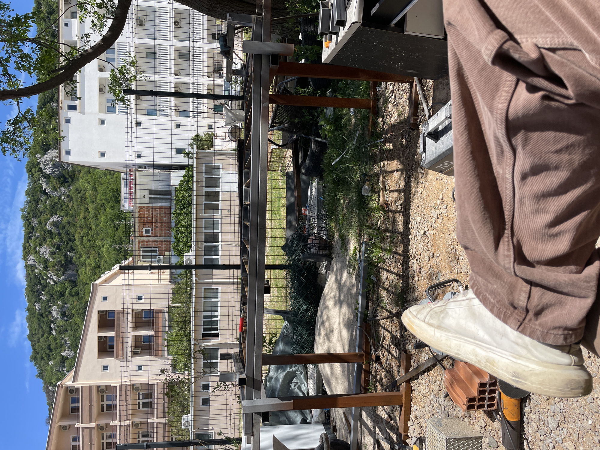 The scene is in Čanj, Montenegro, likely a backyard or construction area. A brown-clad person wearing a white shoe stands on a brick with an angle grinder. Behind them are partially constructed metal frames. Beyond the frames is a wire fence, behind which are residential buildings on a hillside with a mix of green vegetation and rock formations. The sky is blue.
