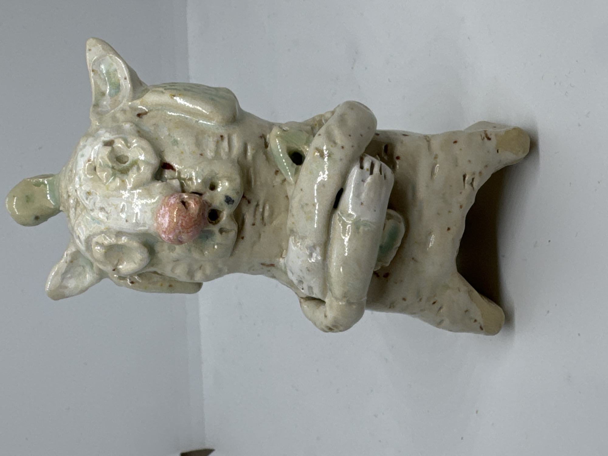 An abstract, cream-colored ceramic sculpture of an animal stands upright against a white background. The sculpture has large, prominent ears, a round pink nose, and what appears to be two eyes formed by circular indentations. Its arms are wrapped around its body, and it clutches a small, light green object. The sculpture has a textured surface with small brown speckles. The lighting is soft, casting a gentle shadow behind the object. The image is a close-up, head-on shot of the sculpture. There is no indication of the city of Waterford, Ireland in the image.