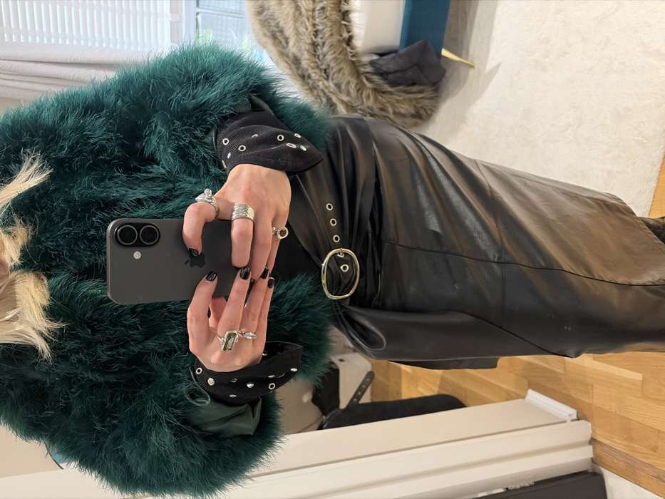 The image depicts an individual indoors, capturing a self-portrait reflection. The exact location within London, United Kingdom, cannot be determined from the visible elements.
A person, with blonde hair, is partially visible from the upper chest to the upper thighs. They are wearing a dark green feathered or faux fur jacket over a dark garment with long, ribbed sleeves that feature multiple silver-toned eyelets/studs. A black leather-look midi-skirt, possibly a wrap style, is worn, cinched with a wide black belt that has a prominent gold-toned oval buckle and several smaller gold-toned eyelets/grommets. Both hands are manicured with black nail polish. The left hand displays a large, rectangular-cut stone ring with a hanging silver-toned accent, along with other silver-toned band rings. The right hand also features multiple silver-toned rings. The individual is holding a black Apple iPhone, model identifiable by its dual rear camera system, indicating the act of taking a photograph, likely a self-portrait via a mirror.
The environment is an interior space, characterized by a light-colored wooden floor on the left, transitioning to a light, shaggy rug or carpet on the right. A light-colored wall is visible to the left. In the background on the right, a window with white horizontal blinds is present. A piece of furniture, possibly a bed or bench, upholstered in teal/blue with gold-toned legs, is partially visible on the far right, covered by a brown, furry throw. The presence of a mirror is implied by the framing of the self-portrait.