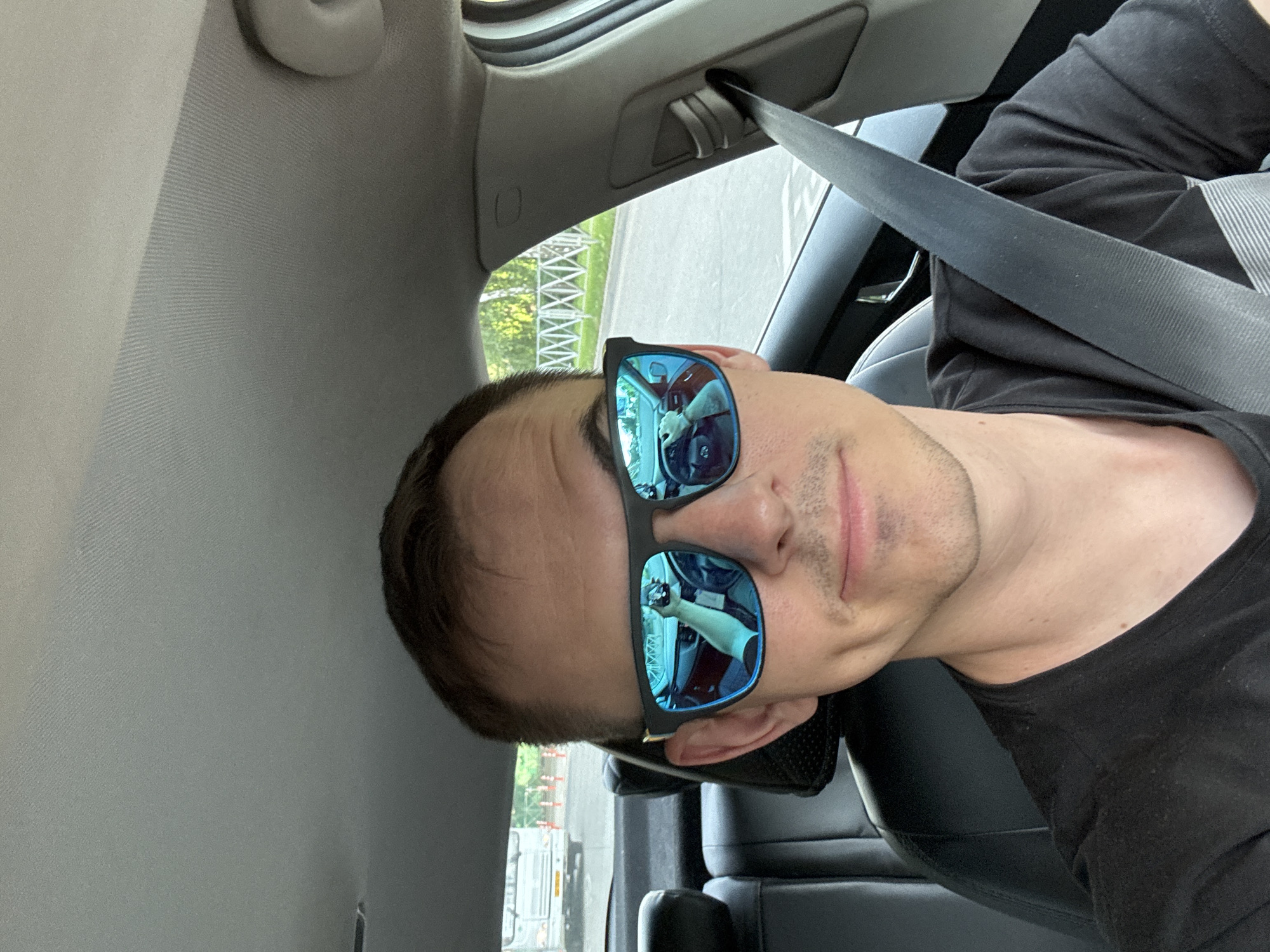 A man is wearing a black t-shirt and blue sunglasses in the driver's seat of a car. He is wearing a seatbelt. The reflection in his sunglasses shows a white dog inside the car and the man holding his phone to take the picture. The background shows a structure of power lines and trees, which could possibly place the location near the intersection of Kalinina Street and Dzerzhinsky Street in Krasnodar, Russia, where similar structures are present.