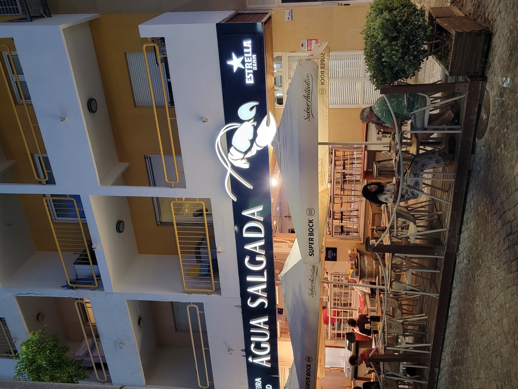 The image depicts the exterior of a restaurant named "Água Salgada" in Quarteira, Portugal. The restaurant features a prominent sign with the name in large white lettering against a dark background, accompanied by a white illuminated lobster graphic. To the right of the lobster is a sign for "Estrella Damm" beer.
The restaurant has an outdoor seating area with several tables and chairs arranged on a wooden deck. White umbrellas, branded with "Super Bock" and "Sabor Autêntico," provide shade over the seating area. Patrons are seated at some of the tables, including a couple in the foreground engaged in conversation. The restaurant appears to be located on a street with a cobbled pavement. The building behind the restaurant has multiple floors with balconies. The overall scene suggests an evening setting, possibly during the warmer months in Portugal.