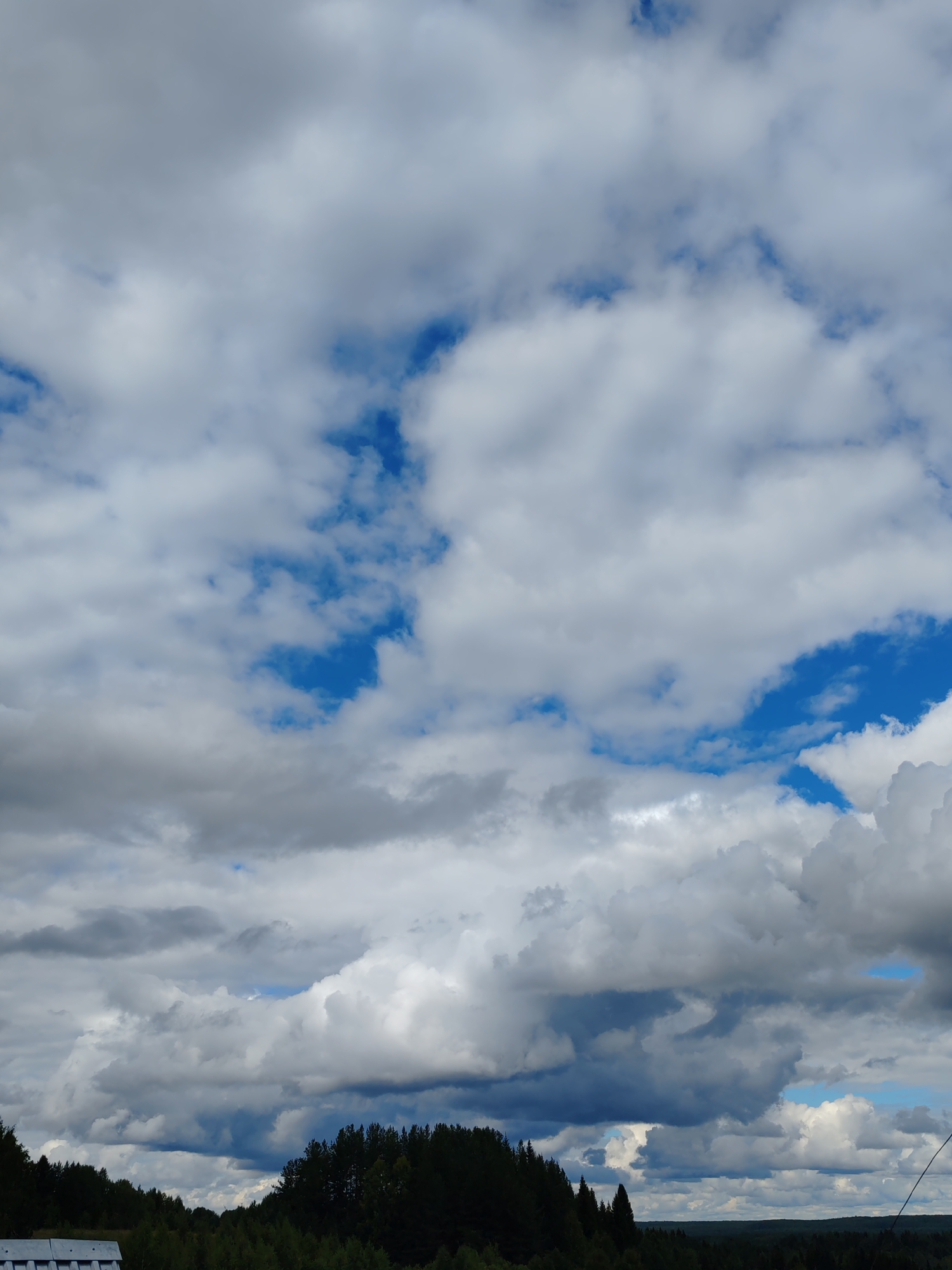 The image depicts a cloudy sky over a dense forest, as viewed from a partially visible rooftop structure in the foreground. The sky is filled with a mix of white and gray cumulus clouds, with patches of bright blue sky visible between them. The forest, a dark green expanse of coniferous trees, stretches across the lower portion of the image. The scene appears to be a natural landscape, likely located in the outskirts of a rural or semi-rural area.