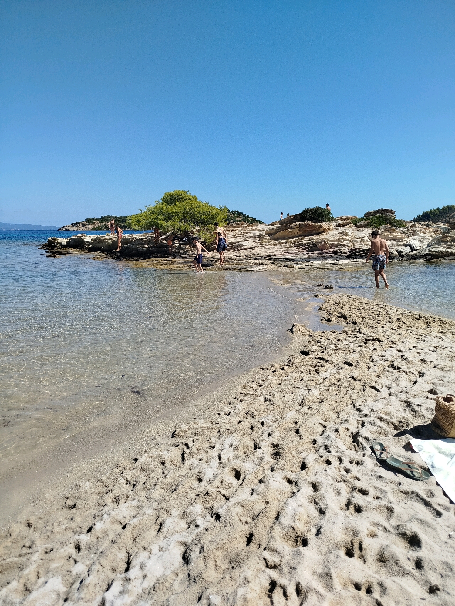 In Sithonia Municipal Unit, Greece, a serene beach scene unfolds under a clear blue sky. The foreground features the textured sandy shore with the gentle lapping of clear, shallow water. A few individuals are scattered across the scene, some wading in the water and others on the rocky shoreline.
To the left, a group of people is clustered on the rocks, appearing to be observing the water or each other. A solitary young male is walking in the shallow water towards the right side of the frame, his body partially submerged. Further to the right, another young male is standing on the rocky outcrop, facing away from the camera.
The background is dominated by a rugged coastline with scattered vegetation and a prominent green tree. The water stretches out to a hazy horizon where it meets the sky. On the beach in the foreground, a woven bag, a towel, and a pair of flip-flops rest on the sand, suggesting a relaxed beachgoing activity.