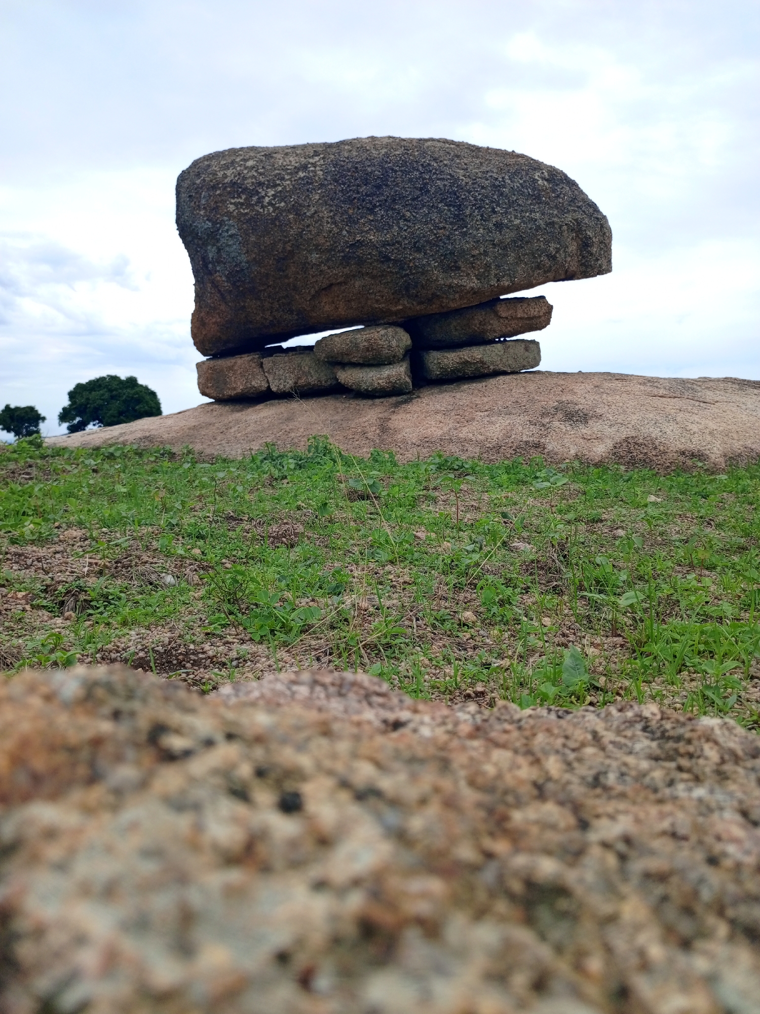 The image displays a natural landscape with a distinctive rock formation. The foreground is dominated by a large, textured rock, out of focus. In the mid-ground, a substantial boulder rests precariously atop several smaller, stacked rocks, creating an unusual, almost balanced structure. This formation sits on a larger, uneven rock surface. The ground around the formation is covered with sparse green vegetation and small pebbles. In the background, two distant trees are visible against a cloudy, overcast sky. The overall scene suggests an outdoor, possibly rural or undeveloped area. The prompt mentions "the city of Jos, Nigeria," but there are no visual cues in the image itself to confirm this specific location.