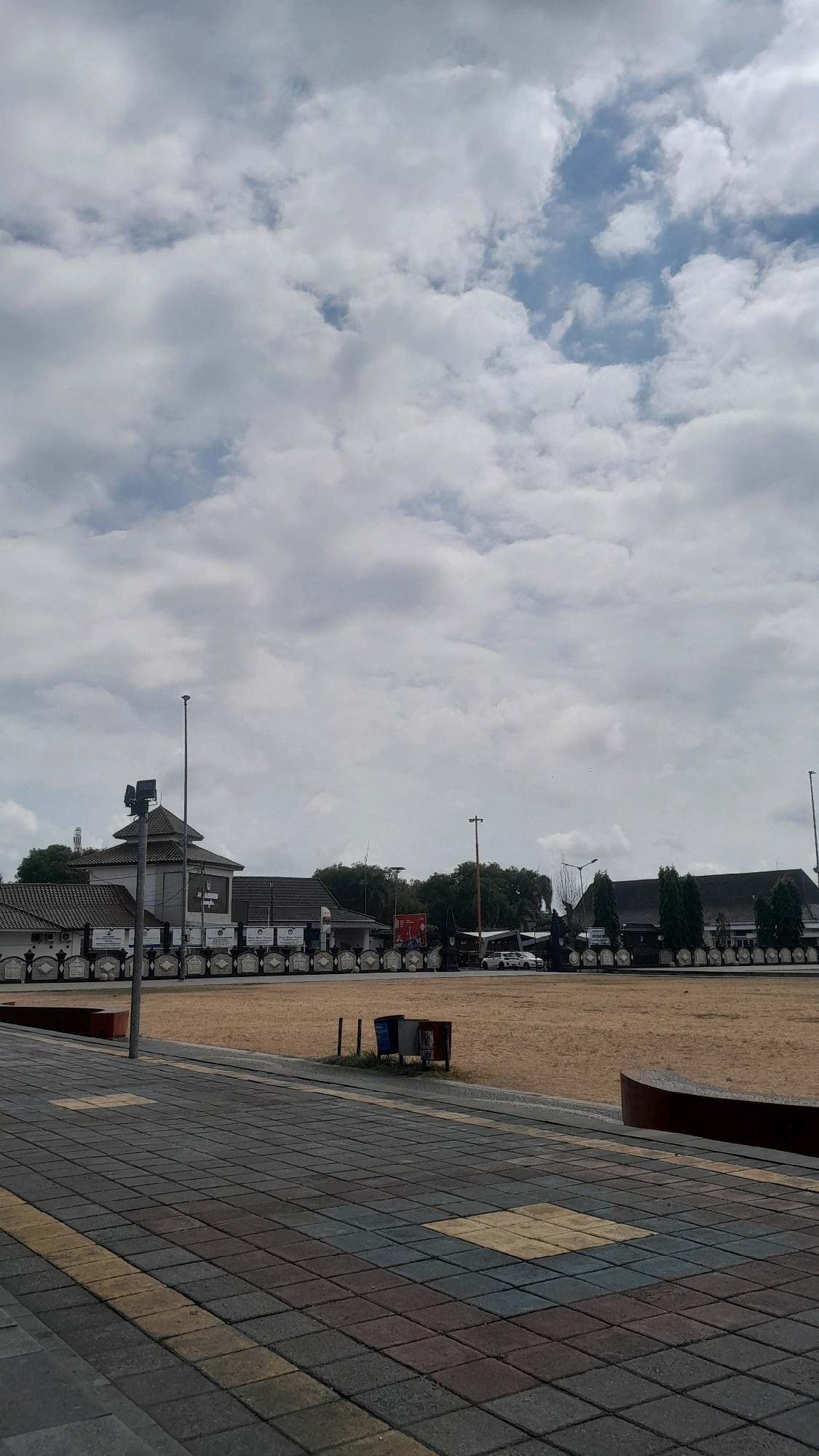 The image captures a daytime scene in the city of Cilacap, Indonesia, likely at a public space or field. The foreground is dominated by a patterned tiled walkway, with alternating colored square tiles in shades of gray, yellow, and reddish-brown.
Beyond the walkway, a large, open field of dry, tan-colored ground stretches into the midground. A low, dark red curved structure is partially visible on the right side of the frame, bordering the field. Two trash receptacles, one blue and one red, are situated near the edge of the field.
In the background, a row of buildings with dark tiled roofs is visible. The architecture suggests a series of connected structures, possibly a complex or institution. A tall, gray pole with a spotlight attachment stands near a building on the left side. Another, thinner pole with a light fixture is visible further to the right. A line of decorative white elements, resembling plaques or individual displays, runs along the front of the buildings. Trees are sparsely planted behind the buildings.
A white car and part of another vehicle are visible in front of the buildings on the right. The sky is filled with a mix of white, fluffy clouds against patches of pale blue. The overall impression is one of an open, perhaps event-ready, public area under a cloudy sky.