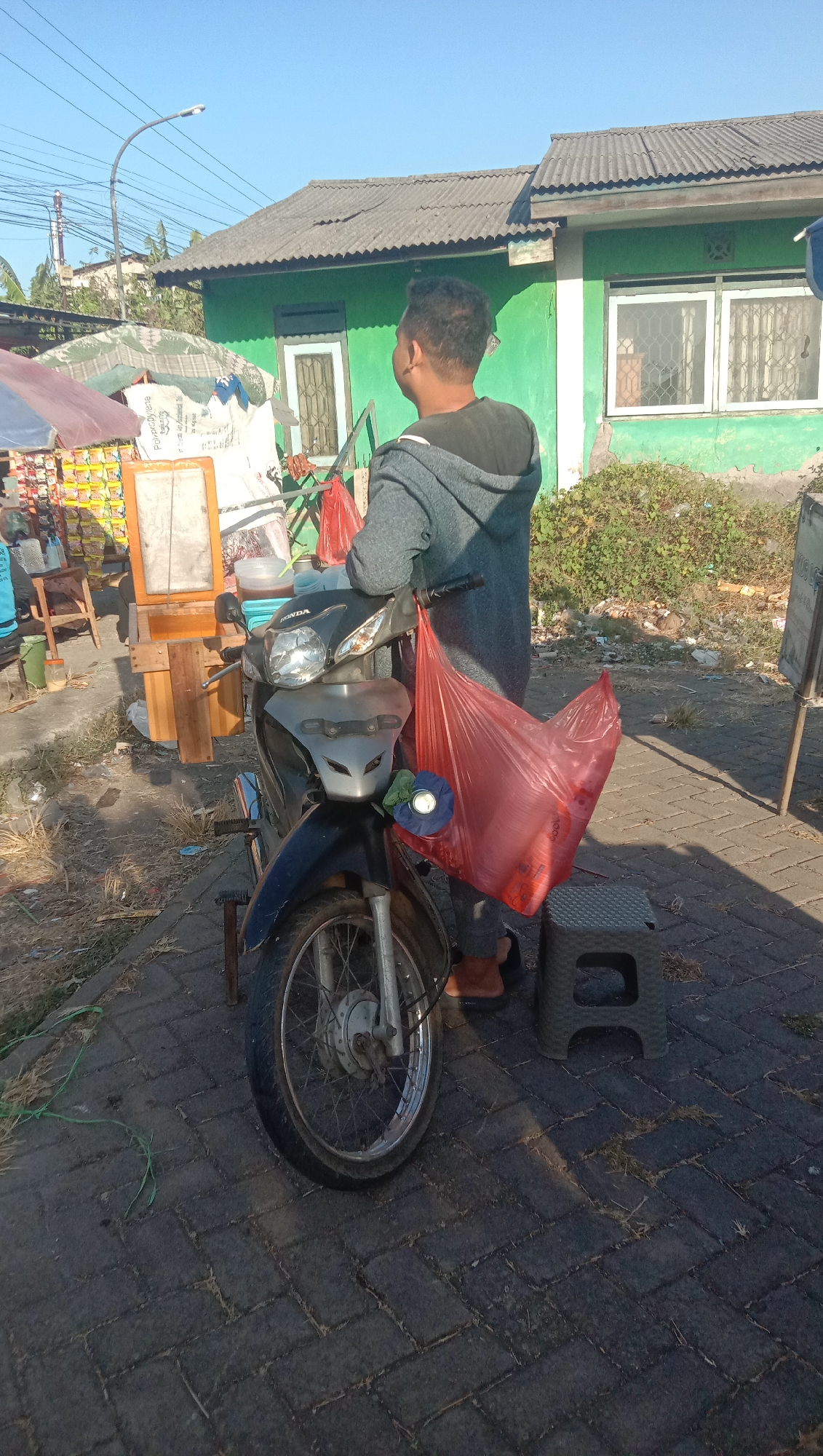 The image depicts a street scene in Curah Dukuh, Indonesia, under a clear blue sky. A man is standing next to a motorcycle, facing away from the camera. He is wearing a gray hooded jacket and dark pants, with brown sandals. The motorcycle, a Honda, has a large red plastic bag hanging from its handlebars, filled with what appear to be plastic cups. Beside the motorcycle is a small, dark gray plastic stool.
In the background, a bright green building with white-trimmed windows stands out. To the left of the building, a stall or cart is visible, laden with various items, including what looks like packaged snacks or food products. A light-colored umbrella shades some of the items on the cart. Electrical wires and a street lamp are present in the upper left of the frame. The ground is paved with dark gray interlocking bricks, with some dry grass and debris scattered around. The overall impression is that of a roadside vendor operating in a residential or commercial area.