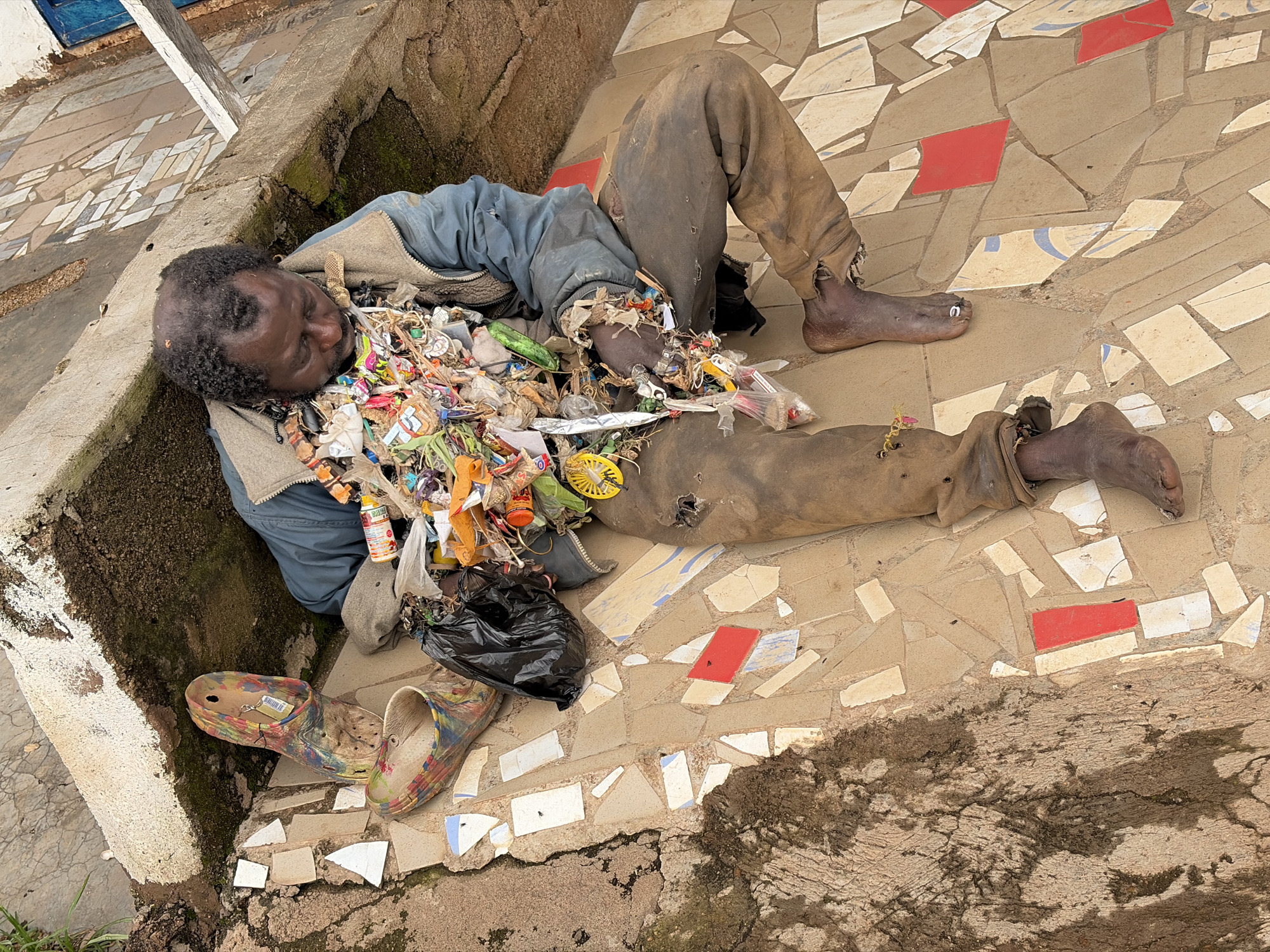 The image depicts a man lying on a mosaic tiled pathway in Jos, Nigeria. He is wearing a blue jacket and dirty brown pants with holes in them. His body is covered in a pile of various types of trash, including plastic wrappers, bottles, and other debris. A black plastic bag filled with trash is also visible next to him. Two colorful clogs are placed near his feet. The man's face is partially obscured by the trash. The background shows a concrete wall and more mosaic tiling. The scene suggests an environment of neglect and poverty.