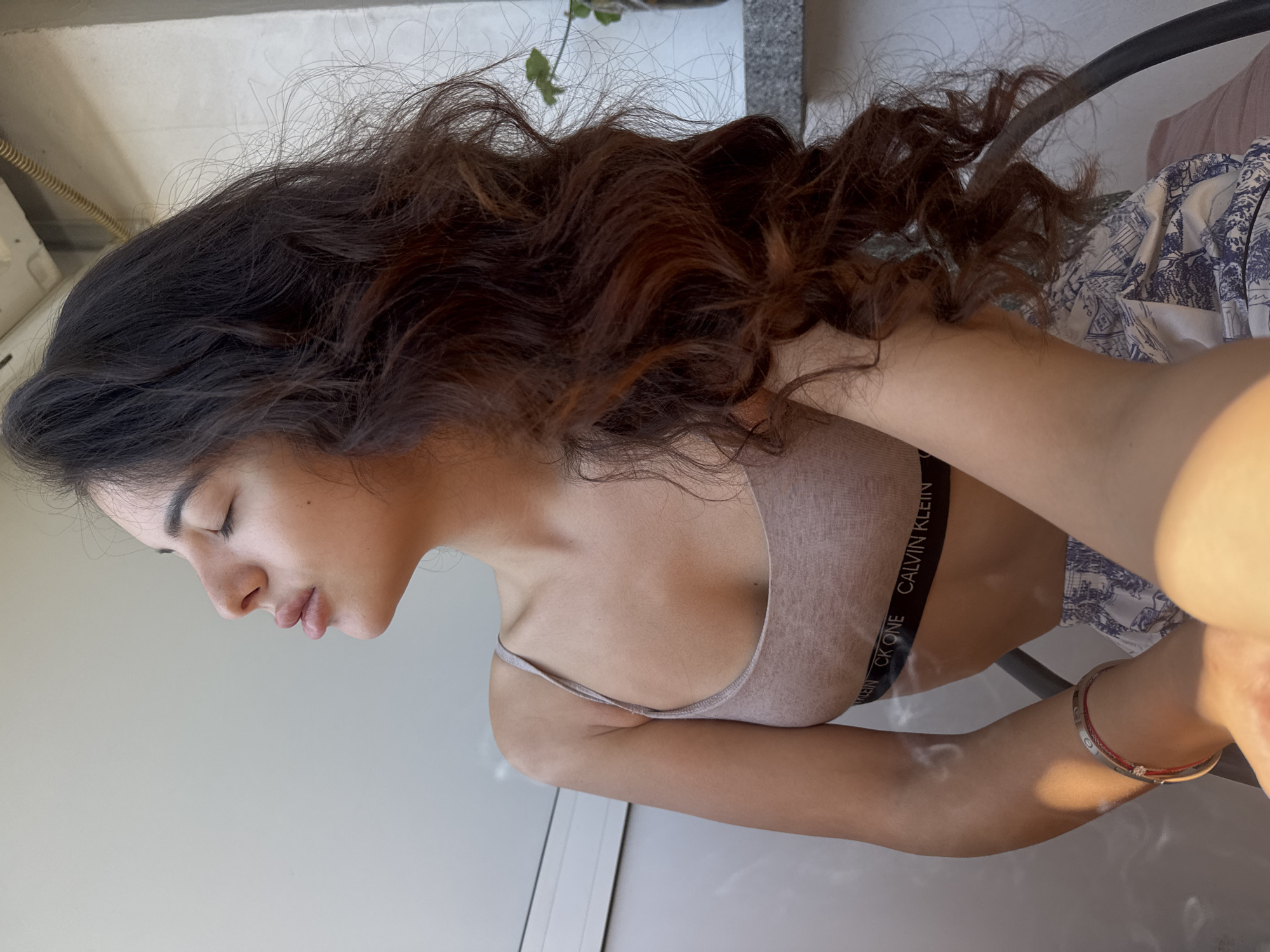 A young woman with long, wavy, dark brown hair is shown from the chest up, seated and facing right with her eyes closed and head tilted slightly back. She is wearing a light pink bralette with a black band that reads "CK ONE CALVIN KLEIN" and patterned shorts. Her left arm is bent, and she is wearing a silver bracelet with red accents. The background consists of a light-colored wall with a horizontal line and a textured ceiling fixture above, suggesting an indoor setting near a window or balcony. A plant is partially visible to the right, and sunlight illuminates the scene, casting shadows and highlighting the woman's skin. The overall impression is one of relaxation. The specific location in the city of Plovdiv, Bulgaria cannot be determined from the image.