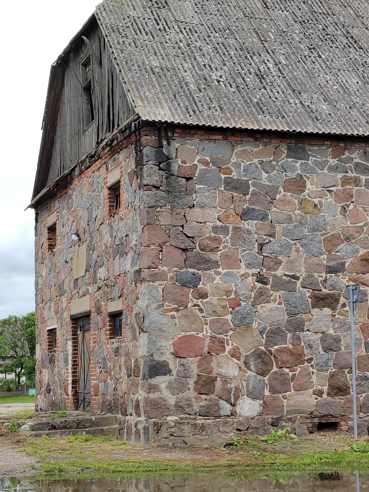 The image shows a weathered, old stone building with a corrugated roof, likely in Воўчынскі сельскі Савет, Belarus. The building's walls are constructed from large, rough-hewn stones of various colors, including gray, red, and brown, interspersed with sections of red brickwork. There are small, dark windows and a set of double wooden doors with metal bracing. The roof appears to be made of corrugated metal or asbestos sheeting, showing signs of age and wear. The building is situated in an outdoor environment with patches of green grass and a puddle of water in the foreground, suggesting it may be damp or recently rained. A gray metal pole or signpost is visible to the right of the building. The sky is overcast. The overall impression is one of an abandoned or disused rural structure.