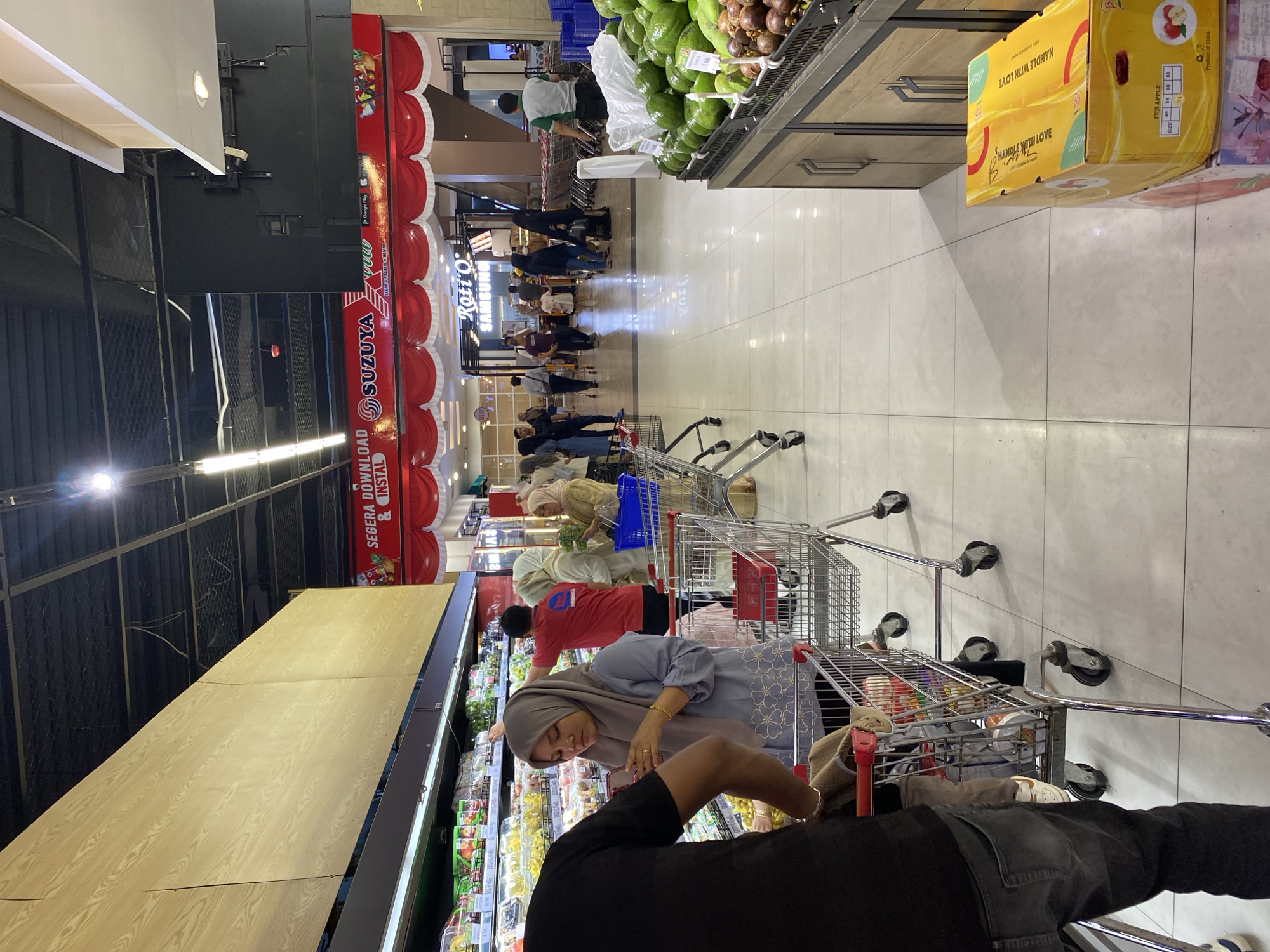 The image shows the interior of a grocery store in Banda Aceh, Indonesia. In the foreground, a person wearing a black t-shirt and grey jeans is pushing a shopping cart. To their right, a woman in a grey hijab and floral patterned blouse is looking at her phone while holding a shopping cart. Behind her, a man in a red t-shirt is reaching for items on a shelf. Further back, several other shoppers and store employees are visible, along with rows of produce, packaged goods, and more shopping carts. A promotional banner with "SUZUYA" and "Roti O" is visible in the background, indicating it is likely a Suzuya supermarket. The store has a tiled floor and overhead lighting.