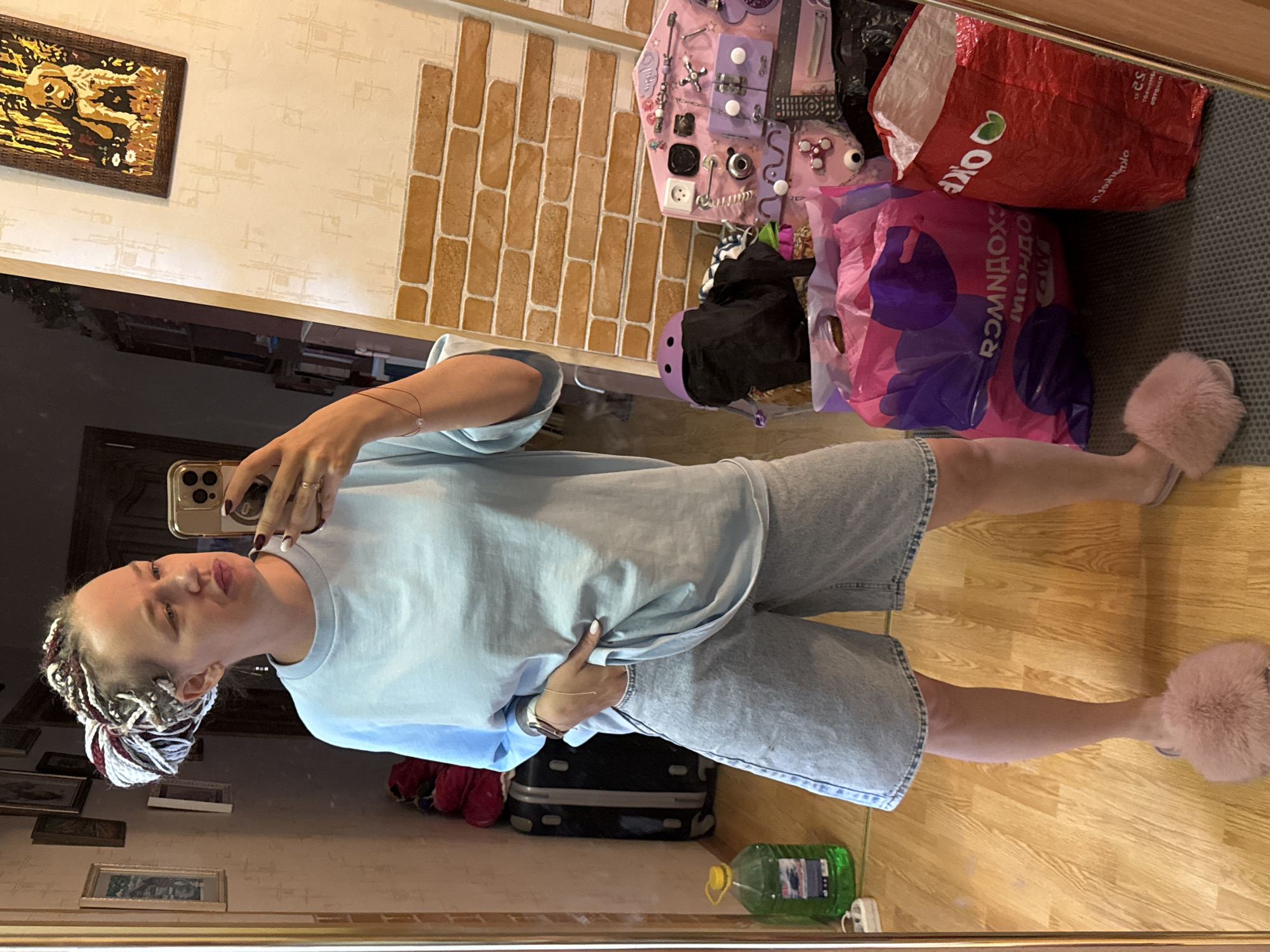 A young woman with braided hair stands in a room, taking a mirror selfie with her phone. She is wearing a light blue oversized t-shirt and denim shorts, paired with fluffy pink slippers. In the background, a large mirror reflects the room, revealing a wall with patterned wallpaper and framed pictures. A suitcase, a large bottle of green liquid, and several shopping bags are visible. A busy toddler's activity board is also present. The room appears to be a domestic setting, possibly a bedroom or a dressing area. The overall impression is of a casual and personal moment captured in time.