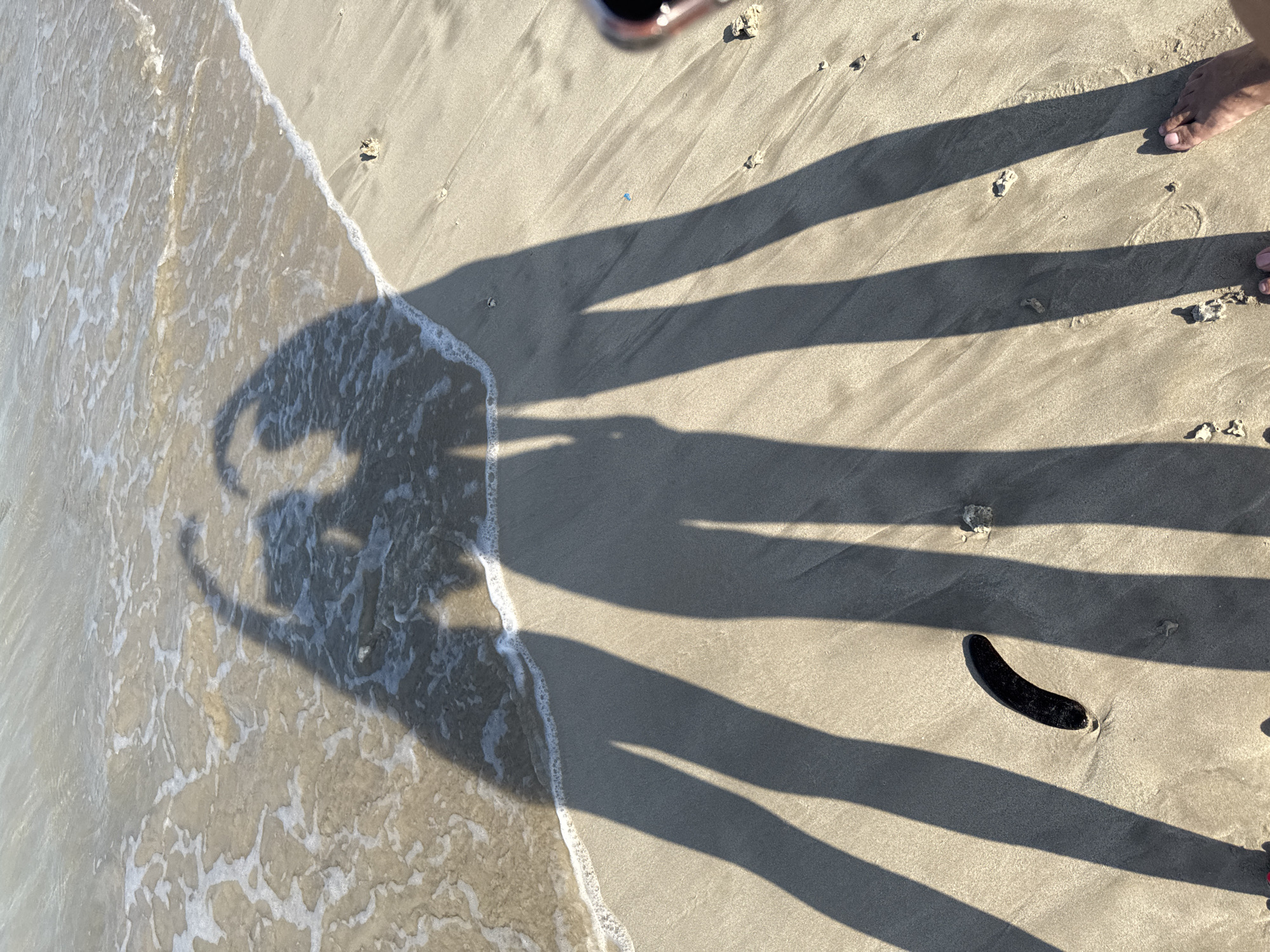 A high-angle shot depicts a sandy beach in Kuta, Indonesia, where ocean waves meet the shore on the left. The shadows of four people are cast onto the sand, and one foot is visible on the upper right. A black object rests on the sand between the shadows of two legs. The sand is speckled with small debris.