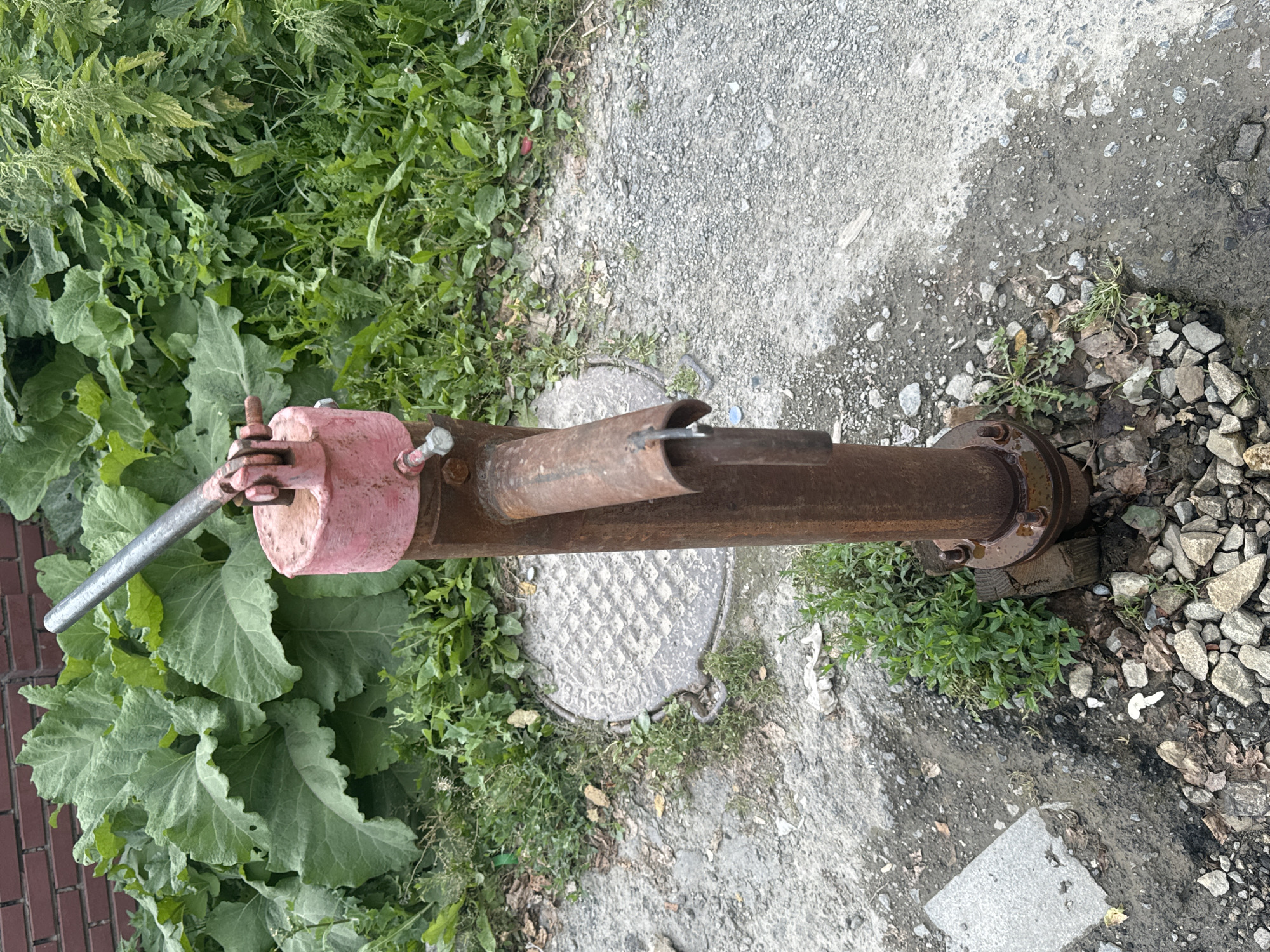 In the city of Unknown, Unknown, a close-up, slightly high-angle shot captures an old, rusty hand pump situated outdoors. The pump, made of weathered metal, stands vertically, its spout directed slightly downwards. A faded pink cap sits atop the pump's mechanism. The ground around the pump is a mix of dirt, gravel, and concrete, with scattered rocks and some patches of green vegetation. Behind the pump, large, green leaves of a leafy plant are visible, and further back, a brick wall is partially obscured by foliage. A metal manhole cover is set into the ground to the left of the pump. The overall scene suggests an outdoor, potentially rural or neglected, urban environment.