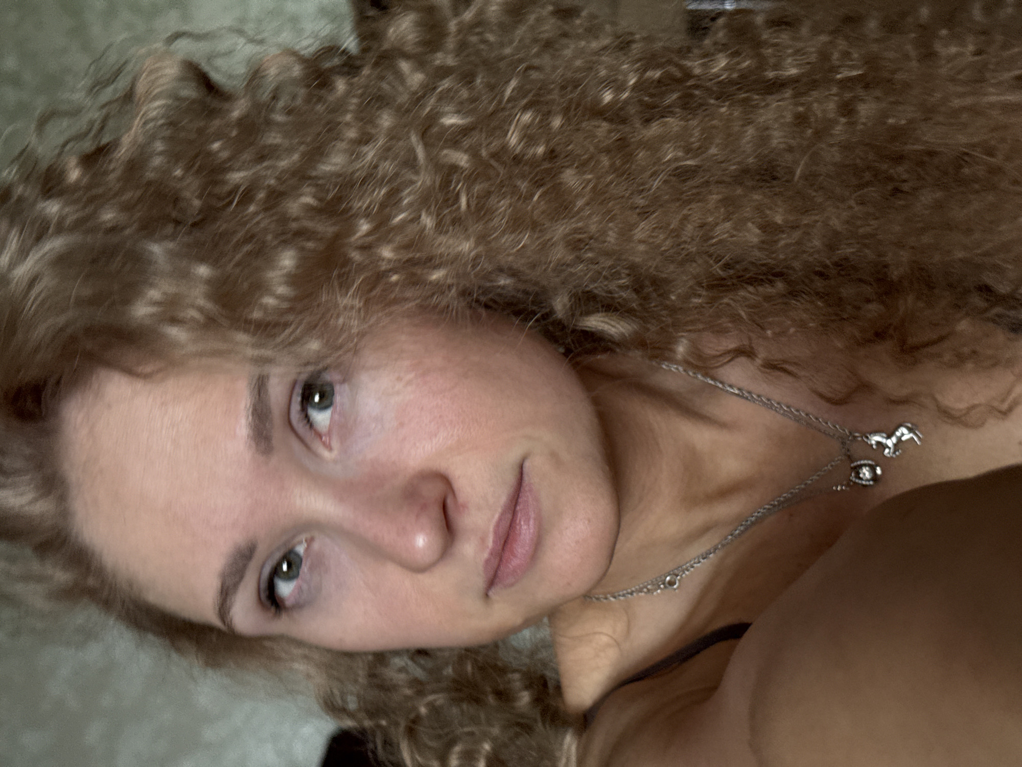 A close-up, slightly angled, portrait-style photograph features a woman with light complexion and curly, brown hair. Her eyes are light green or blue, and she wears a delicate silver necklace with charms. The woman is indoors. There is no clear evidence from the image to support a definitive identification of the city of Perm, Russia.
