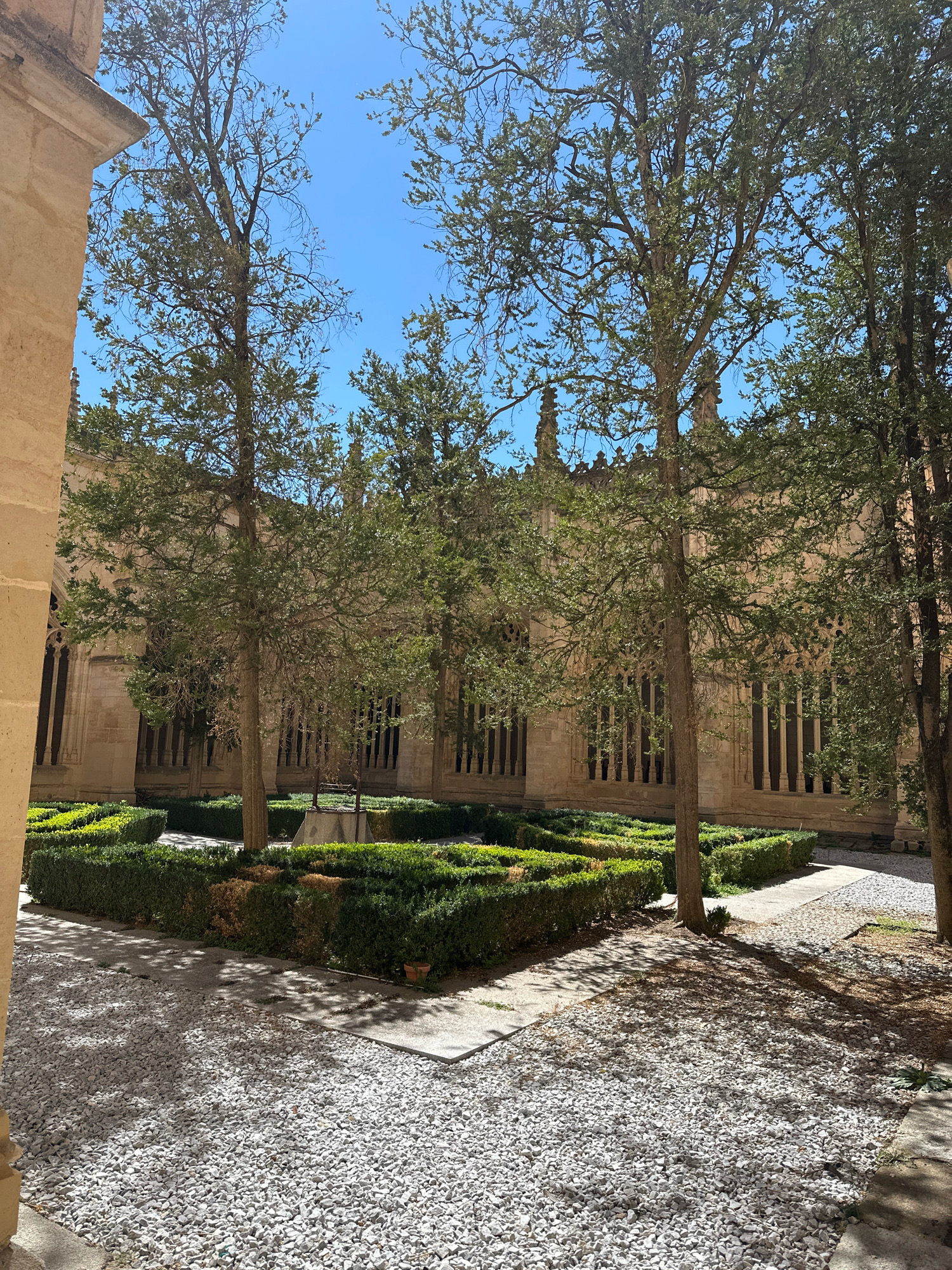 This is an outdoor image of a courtyard with a formal garden and a large building. The courtyard is paved with light-colored gravel and flagstones. In the center of the courtyard is a geometric garden enclosed by neatly trimmed hedges. The hedges form a maze-like pattern with some areas of brighter green foliage. Within the garden, a stone well structure is visible. Several trees with thin trunks and sparse green foliage are scattered throughout the courtyard, casting shadows on the ground. The background features a large, old building constructed of light-colored stone. The building has numerous arched windows with intricate tracery and decorative architectural elements along the roofline. The sky is clear and blue. The image appears to have been taken on a sunny day. The location is identified as a cloister or courtyard within a religious or historical building in La Lastrilla, Spain. There are no people or animals visible in the image.