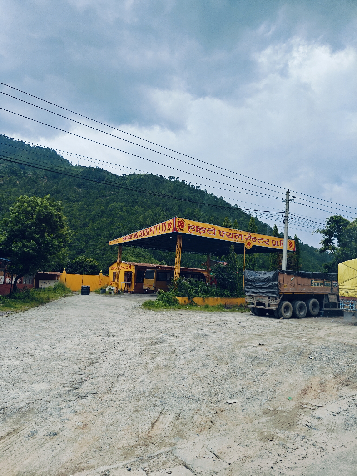 The image depicts a truck refueling at a highway fuel center in Galchhi, Nepal. The fuel center, painted yellow, is situated against a backdrop of lush green hills under a cloudy sky. A large truck with a covered cargo bed is parked at the pumps, its rear wheels visible. Another truck, partially obscured, is parked to the right. The ground in front of the fuel center is a mix of gravel and dirt, showing tire tracks. The signage at the fuel center is in a local script, with "HIGHWAY FUEL CENTER PVT. LTD." also visible in English. Power lines stretch across the upper portion of the image.