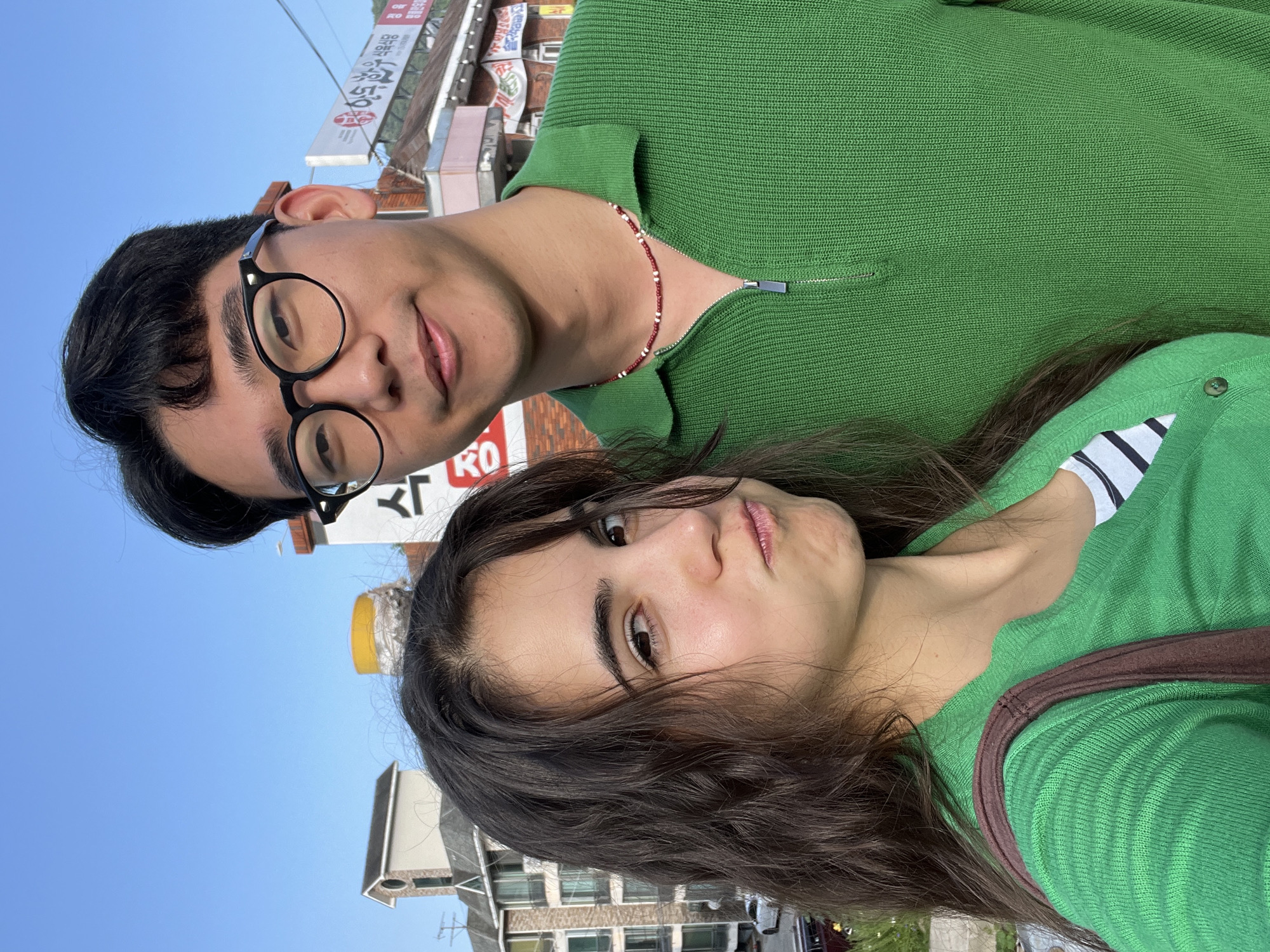 The media file is a close-up, selfie-style portrait of two individuals, a man and a woman, against a backdrop of buildings and a clear blue sky. The image is oriented sideways, as if taken from a phone held vertically.
The man is on the left, wearing black-rimmed glasses and a green collared shirt with a zipper detail. He has dark hair and is smiling gently. A beaded necklace with red and white beads is visible around his neck.
The woman is on the right, with long, dark, wavy hair. She is also wearing a green top, which has a ribbed texture and appears to be a cardigan over a striped shirt. Her expression is neutral, with her lips slightly parted.
The background shows buildings, some with signage that includes Korean characters. One sign clearly reads "RO" in large red letters. There are also hints of streetlights and overhead wires, suggesting an urban or semi-urban environment. The clear blue sky indicates it is daytime, likely during fair weather. The overall impression is a casual, candid moment captured between two people, possibly friends or a couple, in South Korea.
