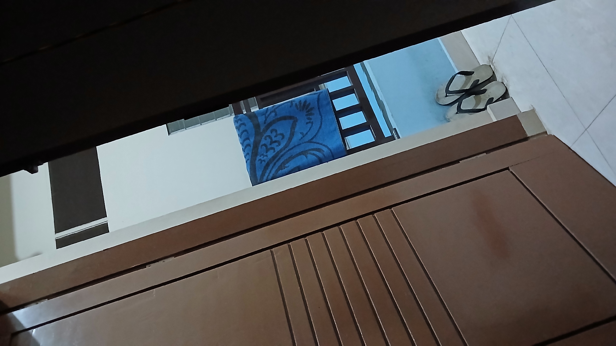 The image shows a view from within a building in Lhokseumawe, Indonesia. A brown cabinet or door is in the foreground. Through a gap above the cabinet, the scene outside reveals a white wall, a blue towel hanging on a wooden rack, and a pair of white flip-flops on the floor near a tiled wall.