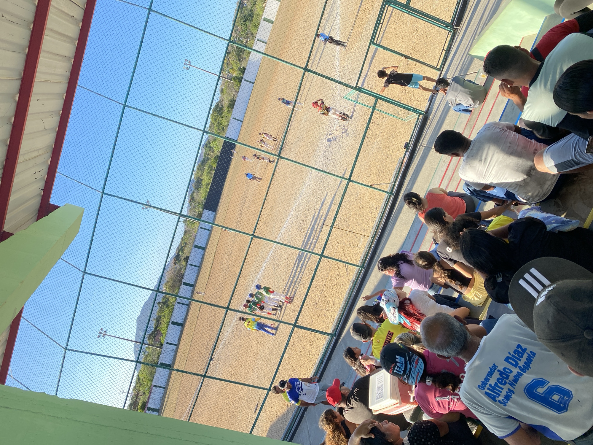 Here is a detailed description of the image:
**Location:** The image shows an outdoor baseball field scene, likely in or near Juan Griego, Venezuela. This is based on the text on a shirt.
**People:** A crowd is gathered in a seating area, potentially a grandstand. Several people are visible in detail. People are sitting and watching the baseball game.
**Subjects:** A baseball game is underway on the field. Several players are visible, in different uniforms.
**Objects:** A chain-link fence separates the grandstand from the baseball field. There are light poles visible in the background of the field. A cooler or container is sitting on the ground.
**Actions:** The players on the field appear to be in the middle of a game, with some near the batter's box and others in the field.
**Interactions:** Spectators are observing the baseball game. The people in the seating area are interacting among themselves.
**Environment:** It is a sunny day with a clear sky. The landscape in the background includes some vegetation.