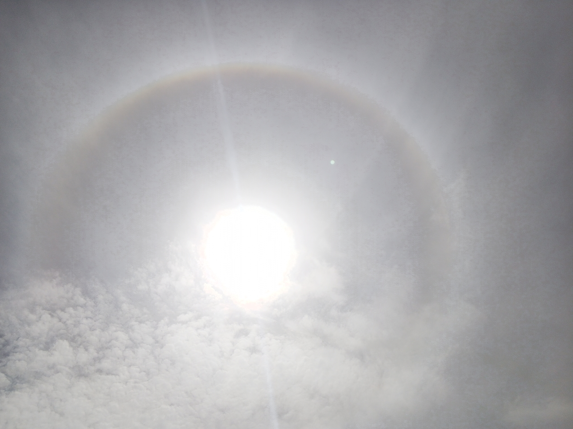 The image shows a sun halo in a cloudy sky. The sun is bright in the center, and a circular rainbow-like halo surrounds it. The clouds are a mix of white and gray, with some appearing lower in the frame. No people, specific locations, or objects other than clouds and the sun are discernible.