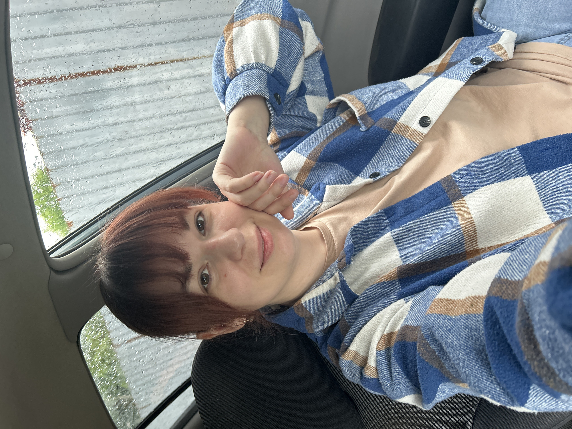 A person with red hair and a blue and white plaid shirt is sitting in a car. Rain is visible on the car window. The location is Фоки, Russia.