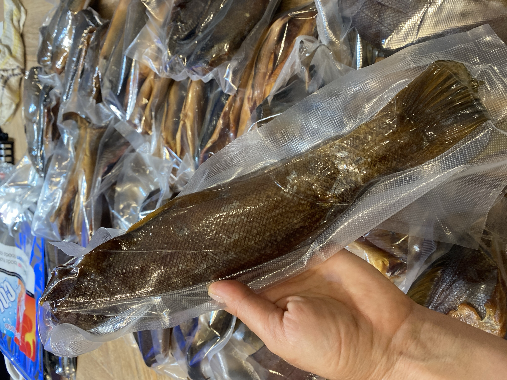 In an indoor setting, a hand holds a vacuum-sealed fish, its scales a mosaic of brown and gold, against a backdrop of more vacuum-sealed fish arranged in rows. The fish is the focal point, its body elongated and tapering towards a fan-like tail. The packaging creates a glossy sheen, reflecting the light and highlighting the texture of the scales. The surrounding fish, also sealed, create a sense of abundance. The image does not provide any visual cues to determine the location in the city of Tret’ya Pad’, Russia.