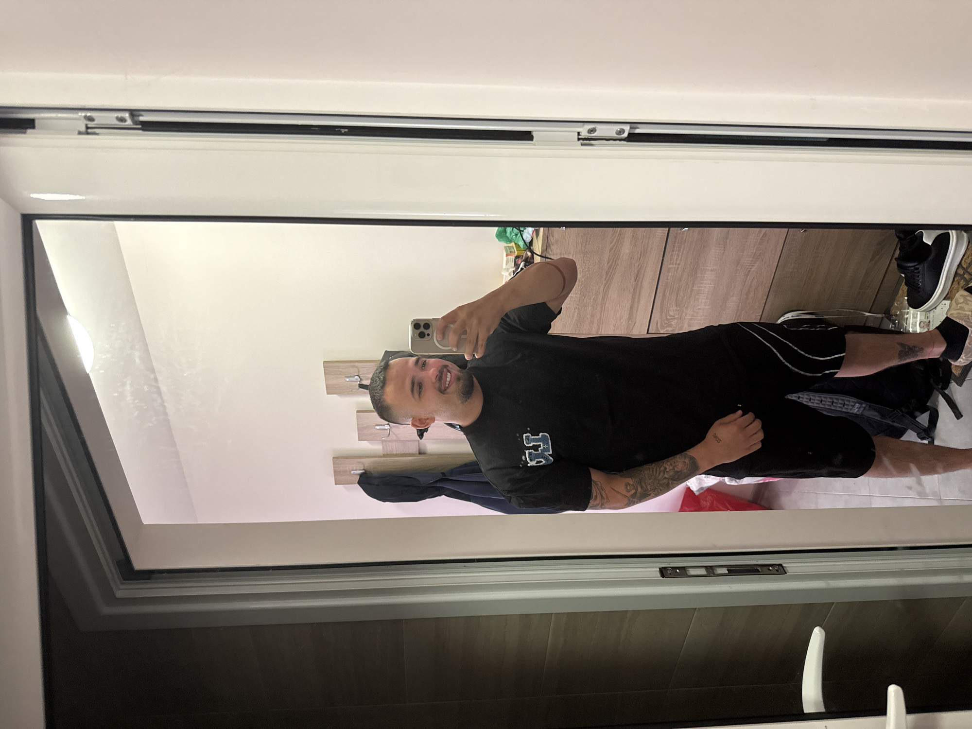 A man is depicted taking a mirror selfie in an indoor setting, likely a bedroom or dressing area in Blagoevgrad, Bulgaria. He is reflected in a large mirror with a white frame and a black inner trim. The man is smiling broadly, holding a light-colored smartphone (possibly an iPhone with multiple camera lenses) with his right hand to capture his reflection.
He is wearing a black short-sleeved t-shirt with partially visible, blocky white lettering on the chest (resembling "LA" or similar), and black shorts with white stripes along the sides. His arms are visibly tattooed; a full sleeve design is on his left arm, and text, possibly reading "deos", is tattooed on his right forearm. He is also wearing black sneakers with thick white soles.
The background reflected in the mirror shows a light-colored wall with a wooden coat rack featuring three wooden panels and silver hooks. To the right, there is a piece of light-colored wooden furniture, possibly a drawer unit or cabinet. The floor reflected in the mirror appears to be light-colored tiles. The top of the mirror structure reveals a white track or sliding mechanism. The overall lighting suggests it is daytime or a brightly lit interior.