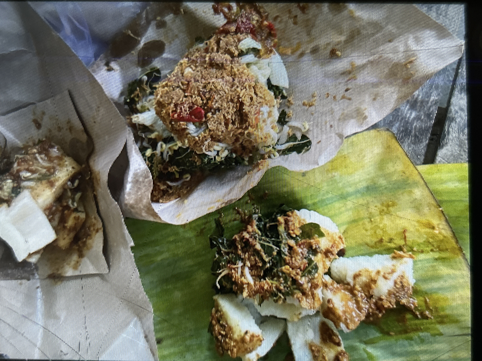 The image depicts a close-up, overhead view of two servings of Indonesian food, possibly nasi campur or similar dishes, presented on banana leaves and paper wrappings. The food consists of rice topped with various ingredients including shredded chicken or fish, vegetables, and a reddish chili sauce. One serving is on the left, partially obscuring a banana leaf, and appears to be composed of several pieces of white rice covered in the toppings. The second serving is to the right, presented in a paper wrapper and also topped with the mixed ingredients. The background shows a textured surface, possibly a wooden table, with some of the food served on white paper. The overall presentation suggests a casual dining setting, possibly a street food stall or a simple eatery in Depok, Indonesia. The lighting is natural, and the image exhibits a slight grainy texture, possibly due to low light conditions or the camera used. No people are visible in the image.
