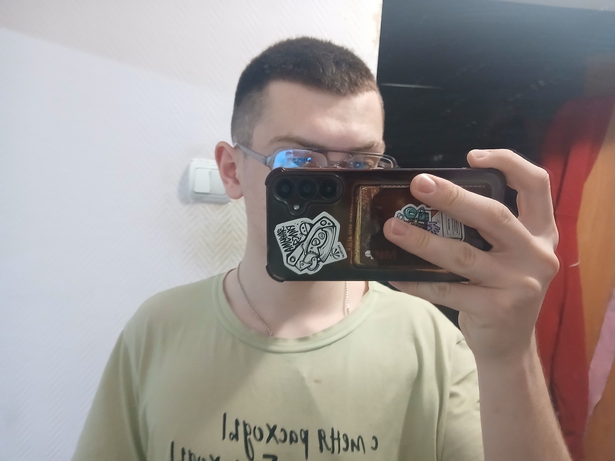 A young man is taking a selfie indoors. He is wearing glasses and a light green t-shirt with writing on it. He is holding a dark-colored smartphone with stickers on the back. The background includes a white wall with a light switch and a darker wall with what appears to be a poster or painting. The location is Chaikovsky, Russia.
