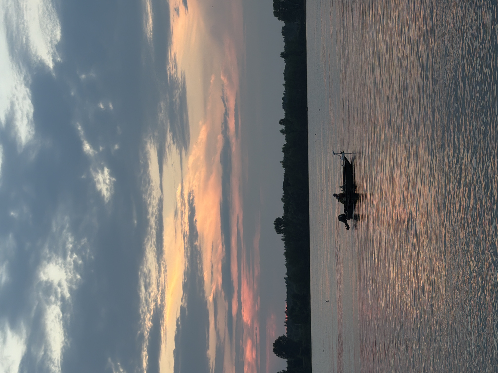 A wide shot captures a tranquil river scene at dusk near Chaikovsky, Russia. The sky is a gradient of muted blues, grays, and soft oranges, with scattered clouds hinting at the setting sun. The water's surface reflects the sky's colors, creating shimmering ripples. In the distance, a dark, tree-lined horizon suggests the presence of a forest. A small fishing boat, silhouetted against the water, is the focal point. A lone figure can be seen in the boat, appearing to be fishing or navigating. The overall atmosphere is one of quiet solitude and natural beauty.