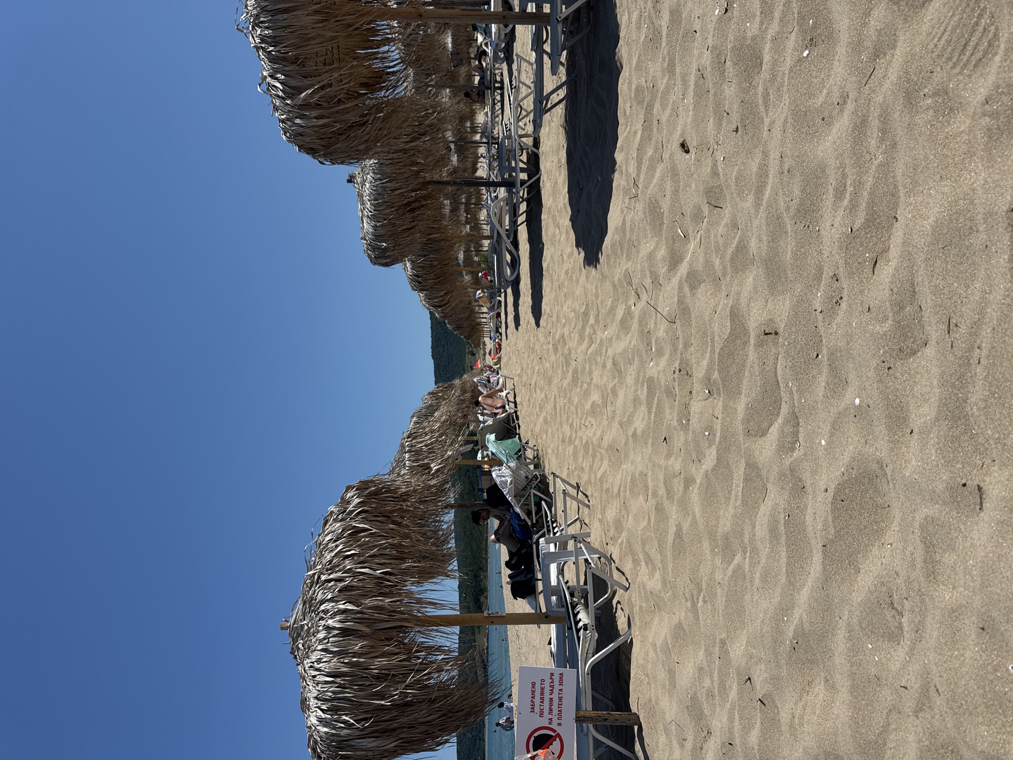 This is a wide shot of a sandy beach on a sunny day, with the Black Sea visible in the background. The beach is lined with rows of thatched umbrellas and empty lounge chairs. Several people are seated on the lounge chairs under the umbrellas, relaxing and looking out at the sea. In the foreground, a sign with a red circle and a diagonal line over an umbrella symbol, accompanied by text in Bulgarian, is visible. The text translates to "It is forbidden to place personal umbrellas in the paid zone." The overall scene depicts a typical resort beach scene in Dyuni, Bulgaria.