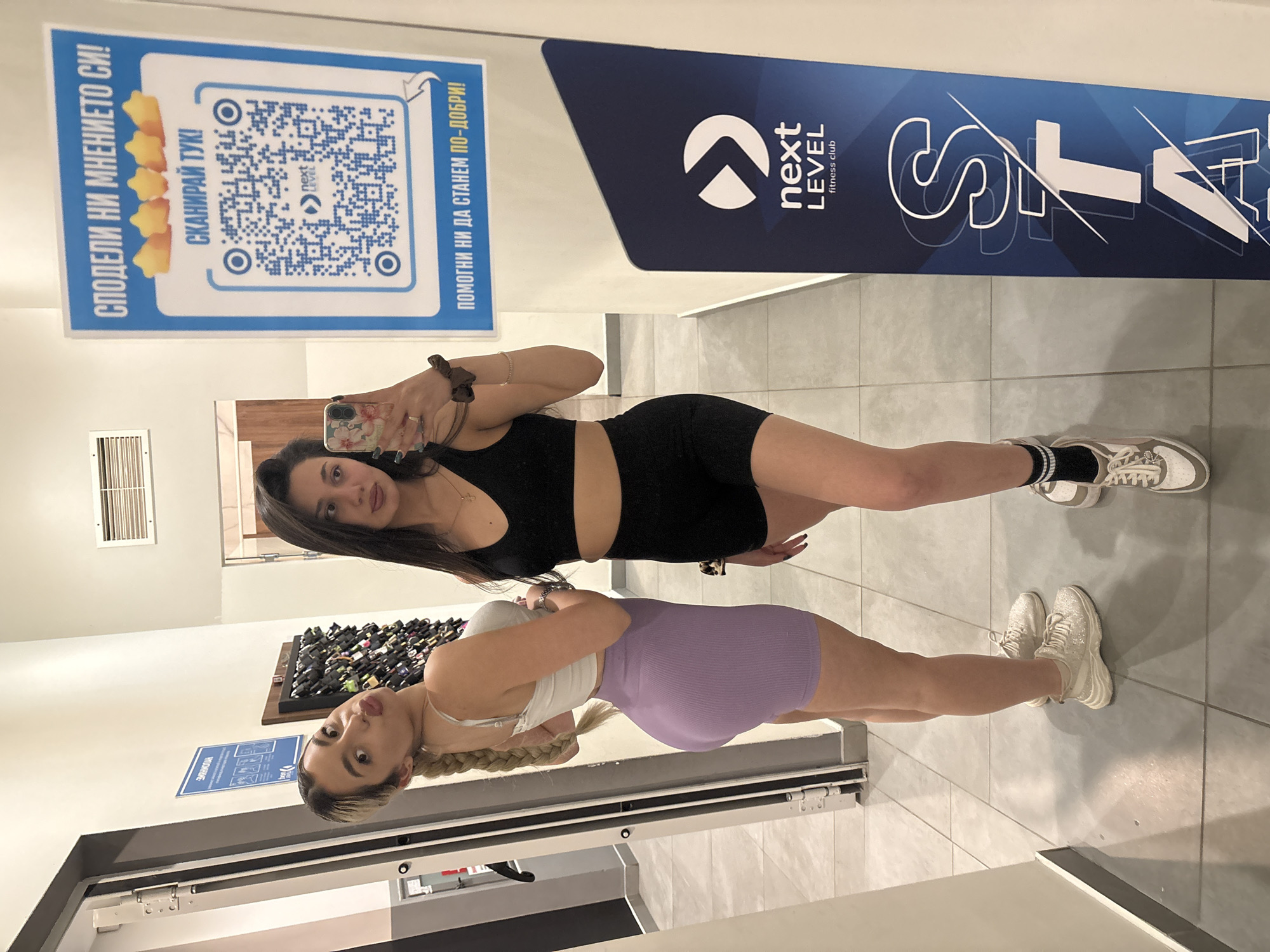 Two young women in workout attire are posing in front of a mirror inside the "Next Level" fitness club. The brunette, on top, is wearing a black crop top and matching shorts, while the blonde, on the bottom, sports a white crop top and a purple ribbed skirt. The club's branding is visible on a poster with the logo. A QR code and additional text in Bulgarian are also displayed on a sign. The setting appears to be a corridor or entryway in the fitness club, with neutral-colored tiles on the floor. A dark display of items (possibly gym equipment) is visible in the background, and an air vent is visible on the wall.