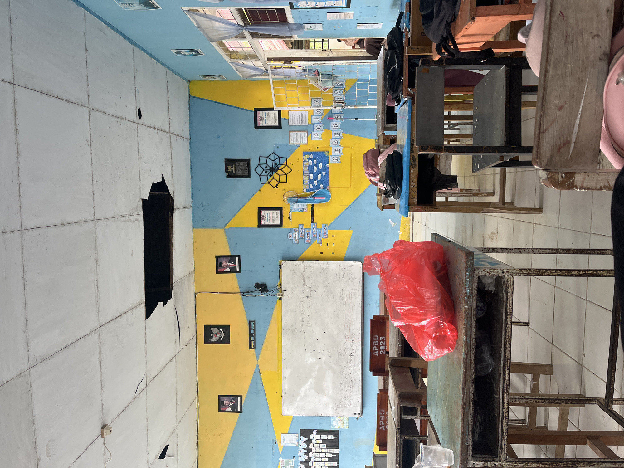The image depicts the interior of a classroom in Pompanua, Indonesia. The room features blue and yellow painted walls with a geometric design. A large whiteboard is mounted on one wall, covered in handwritten notes and diagrams. Several framed pictures of individuals and an emblem are displayed on the walls. The ceiling has visible damage with a large hole. Desks and chairs, some appearing worn and aged, are arranged throughout the room. A bright red plastic bag is placed on one of the desks in the foreground. The floor is tiled. Windows with curtains are visible on one side of the room. In the background, a person can be partially seen through a doorway. The overall environment suggests a functional but somewhat aged educational setting.