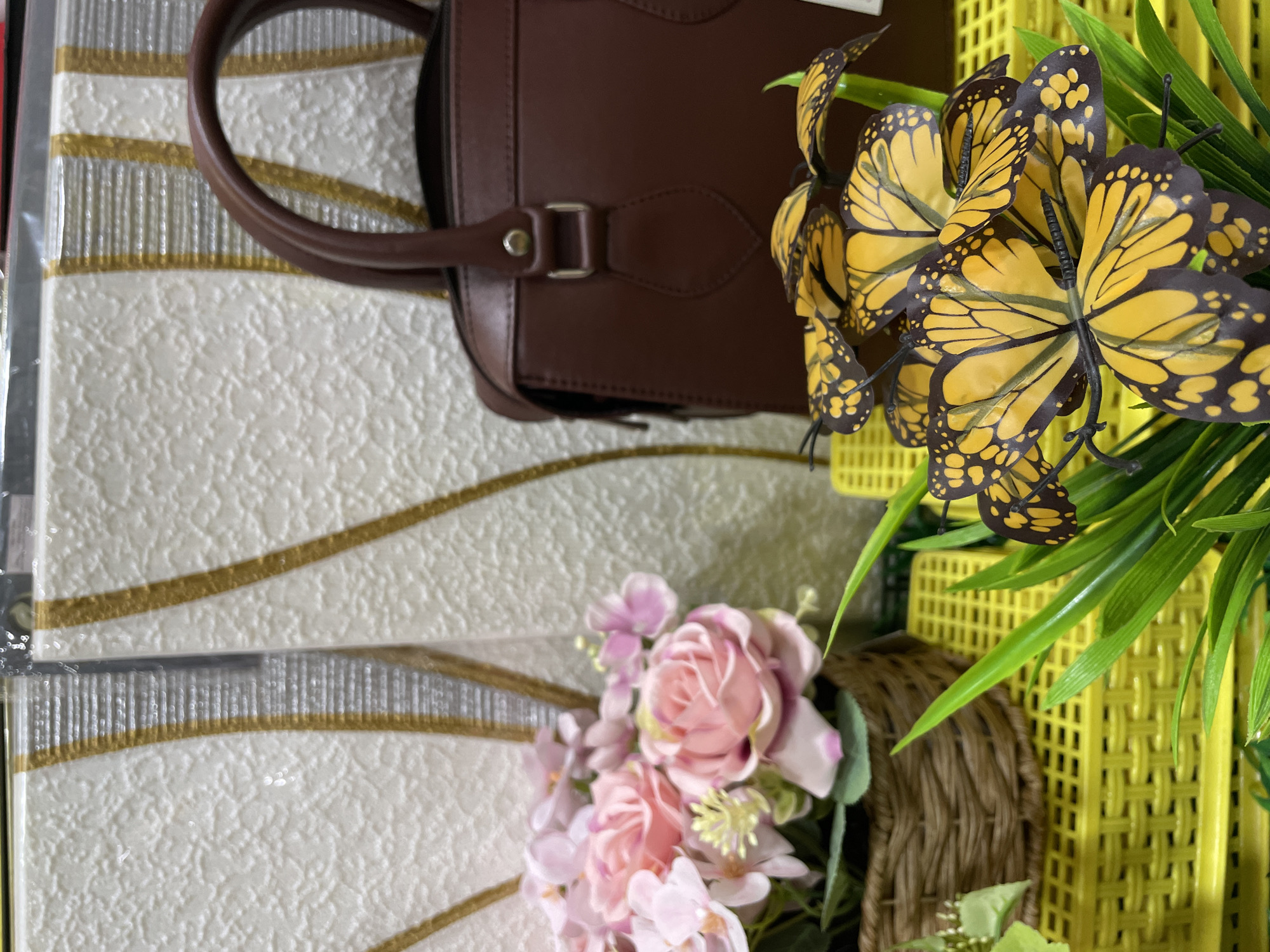 In what appears to be a retail display in Davao City, Philippines, a dark brown handbag with a rounded handle sits prominently. To its left, a woven basket holds a bouquet of artificial pink roses and smaller pink flowers, accented by green leaves. In the foreground, a cluster of artificial yellow and black butterflies, likely decorative, are interspersed with vibrant green artificial foliage. The background features a textured wall with vertical gold stripe accents. The objects are arranged on a yellow, grid-like surface, suggesting a store shelf or display stand.